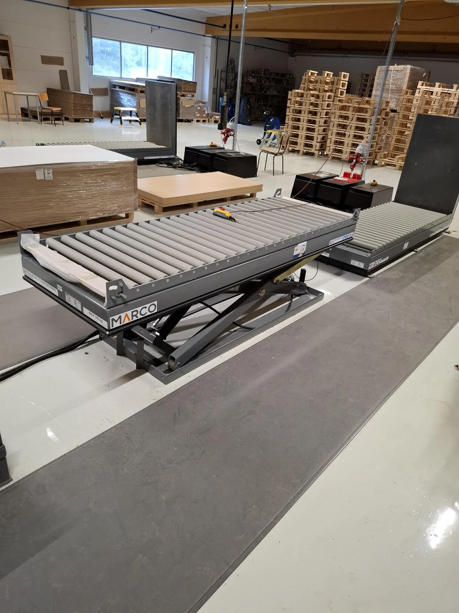 Two conveyor rollers at a furniture warehouse or factory with stacks of wooden pallets and furniture in the background.