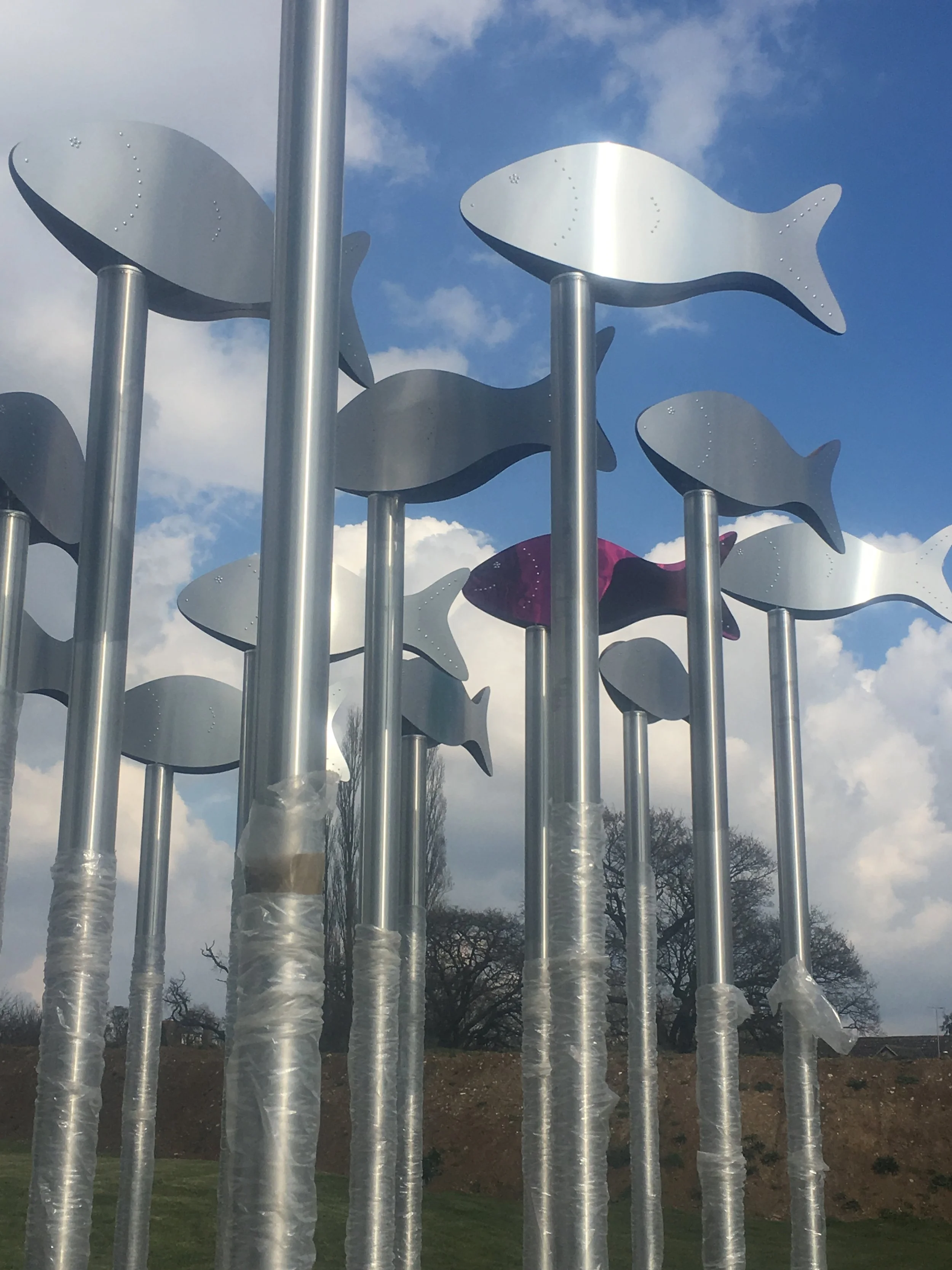 Outdoor sculpture of metallic fish cutouts mounted on tall poles against a cloudy blue sky.