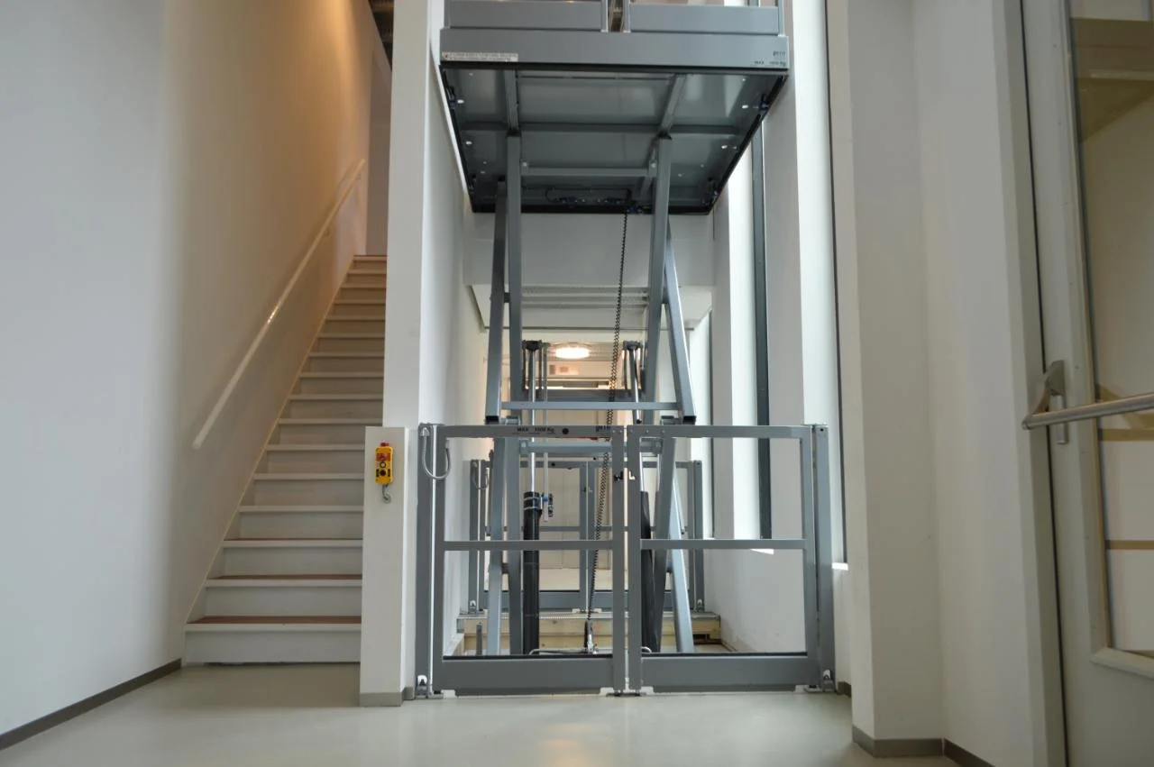 An elevator with a metal frame and an open ceiling, showing its mechanical components, located between a staircase and a wall in a bright indoor setting.