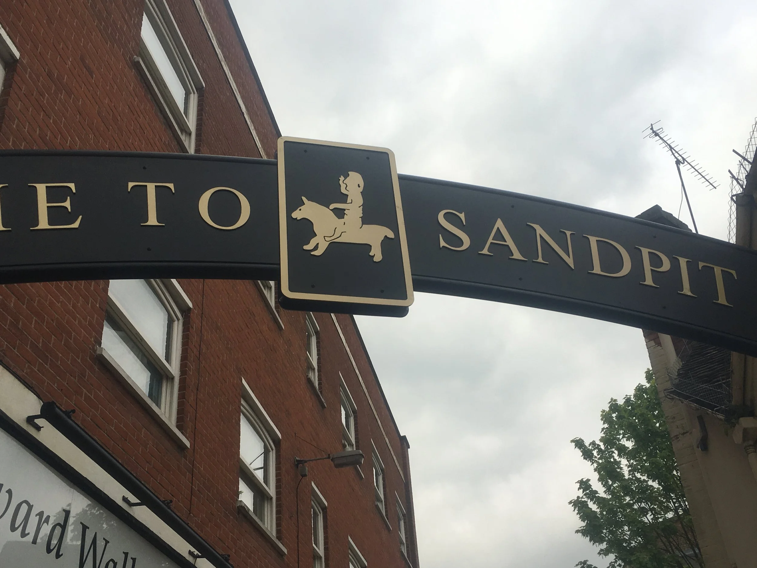 Street sign with the text 'To Sandpit' and a graphic of a girl riding a pig, against a brick building and cloudy sky.