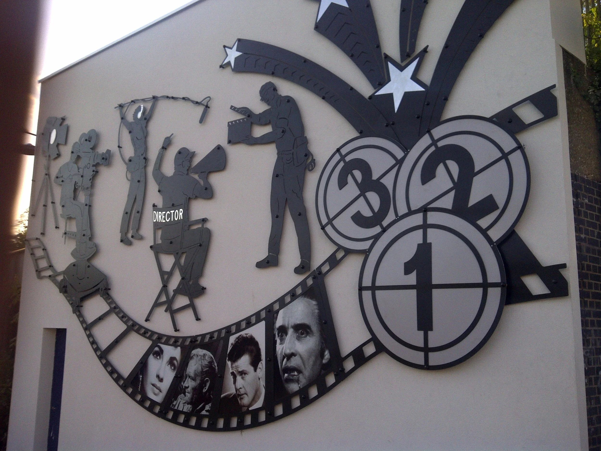 A decorative wall clock featuring silhouettes of film director Alfred Hitchcock, actors, and a film-related theme, with a series of black-and-white photographs at the bottom.
