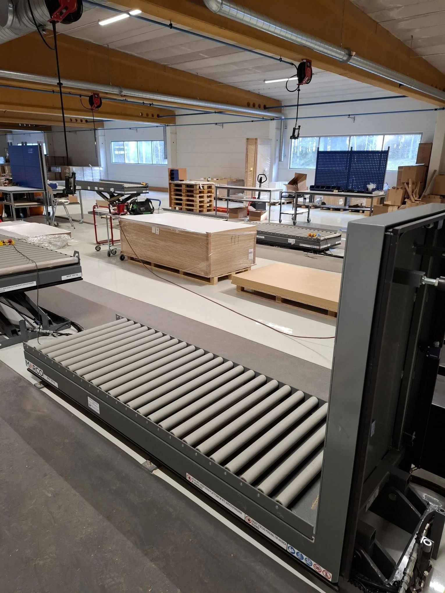 An indoor workshop or manufacturing space with various equipment, conveyor rollers in the foreground, and worktables and boxes in the background, with large windows and overhead lighting.