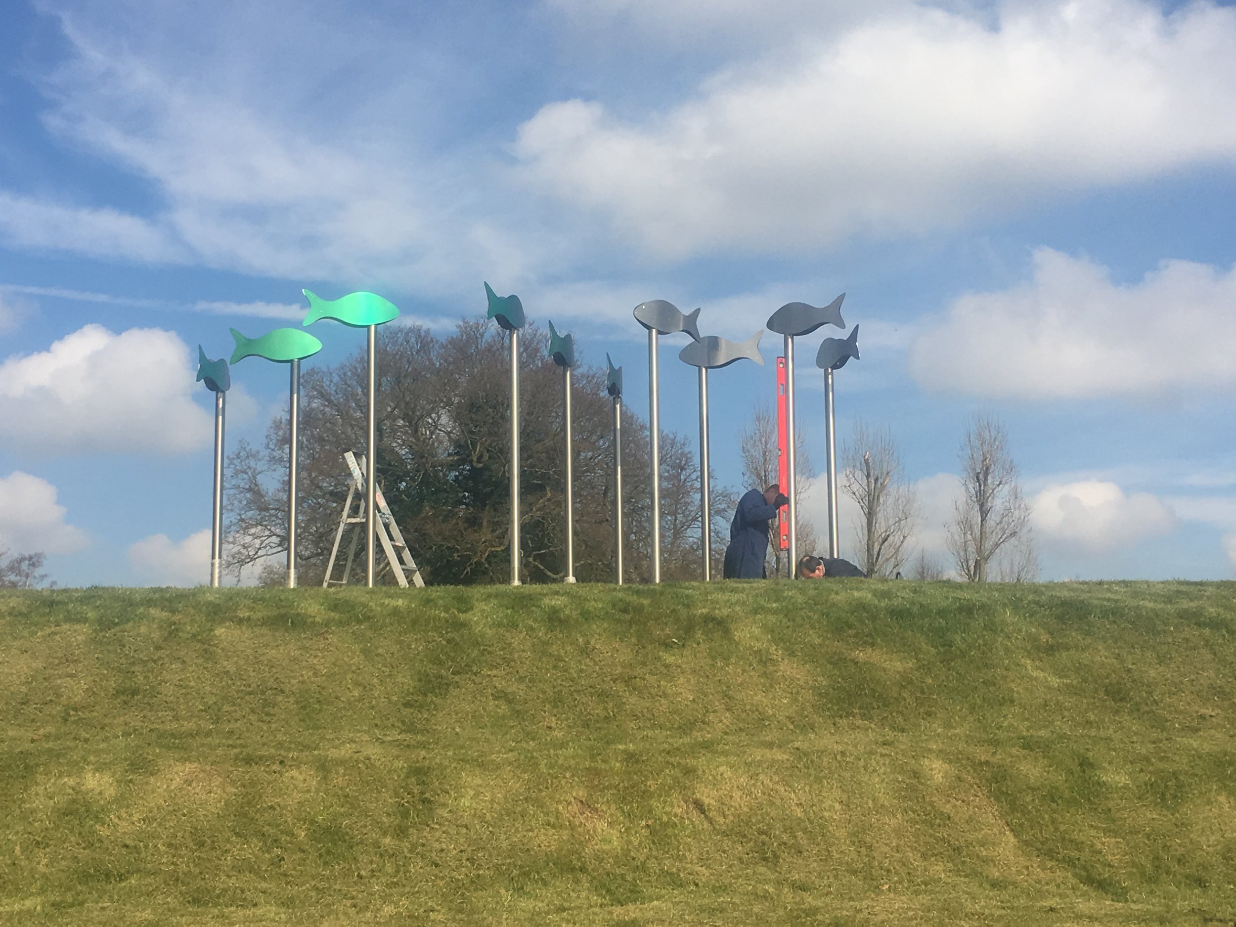 Installation of colorful fish-shaped sculptures on poles on a grassy hill with a blue sky and clouds in the background.