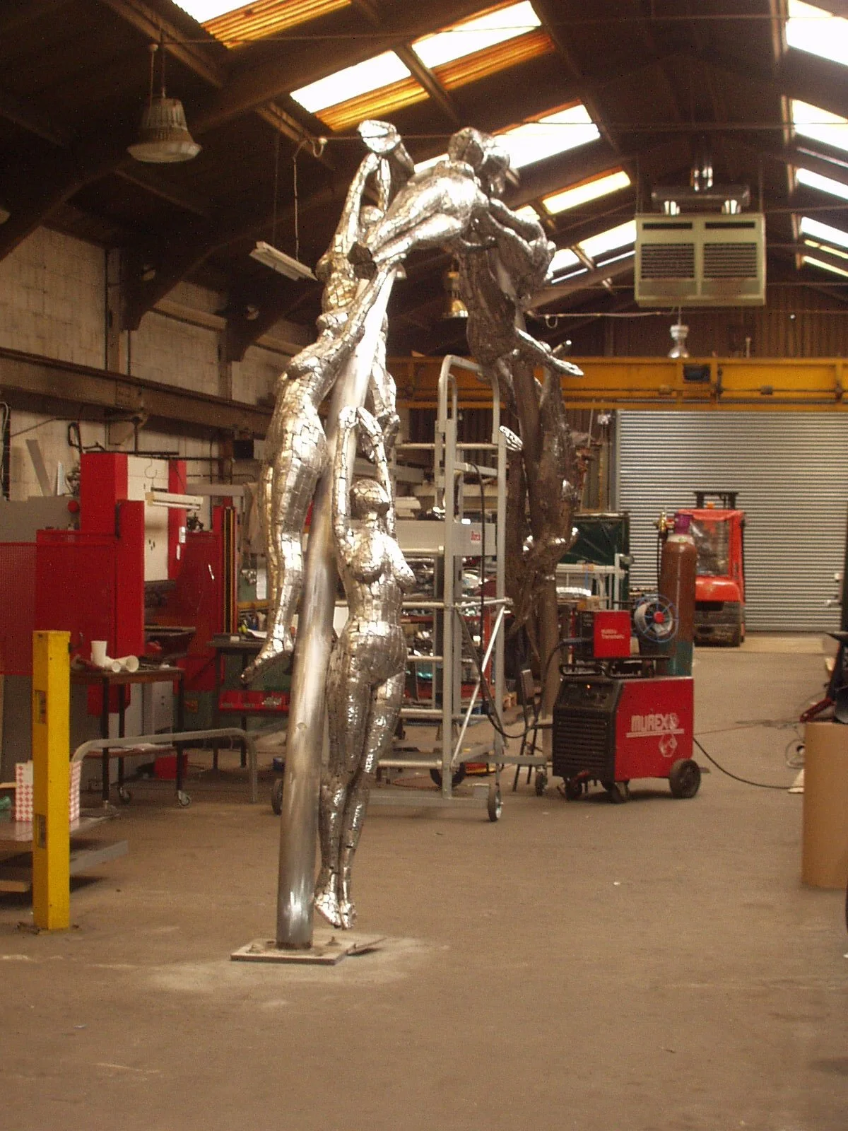 Metal sculpture of human figures in a workshop, with a high ceiling and industrial equipment in the background.