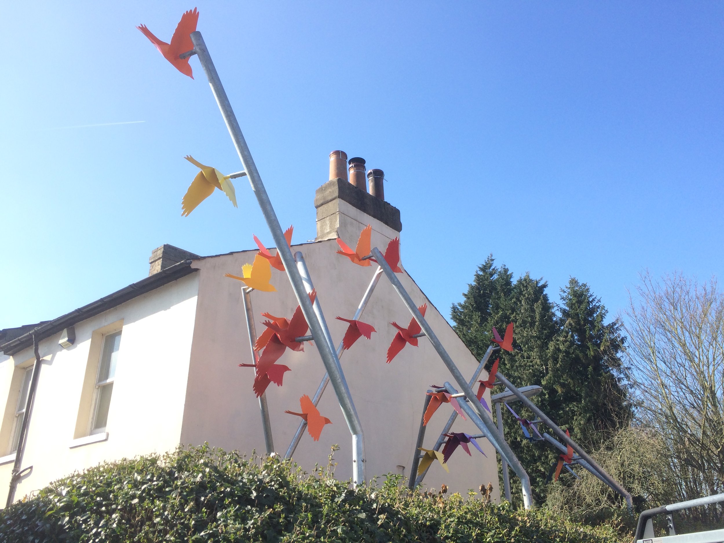 Colorful metal sculptures of birds in orange, yellow, and red perched on metal rods in front of a white house with a chimney and trees in the background.