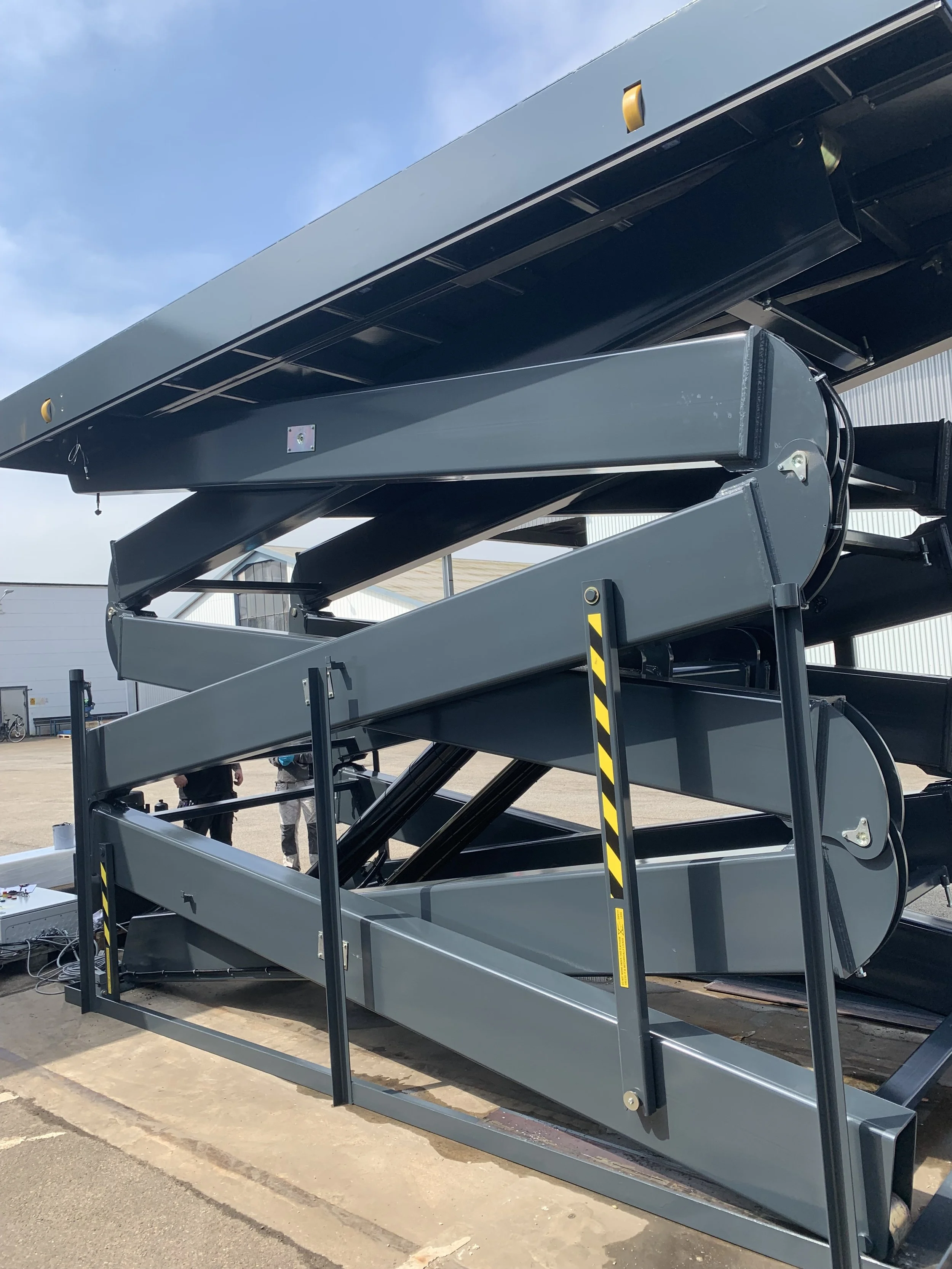 A large industrial scissor lift platform in black and gray, partially extended, outdoors on a paved surface with a blue sky and some industrial buildings in the background.