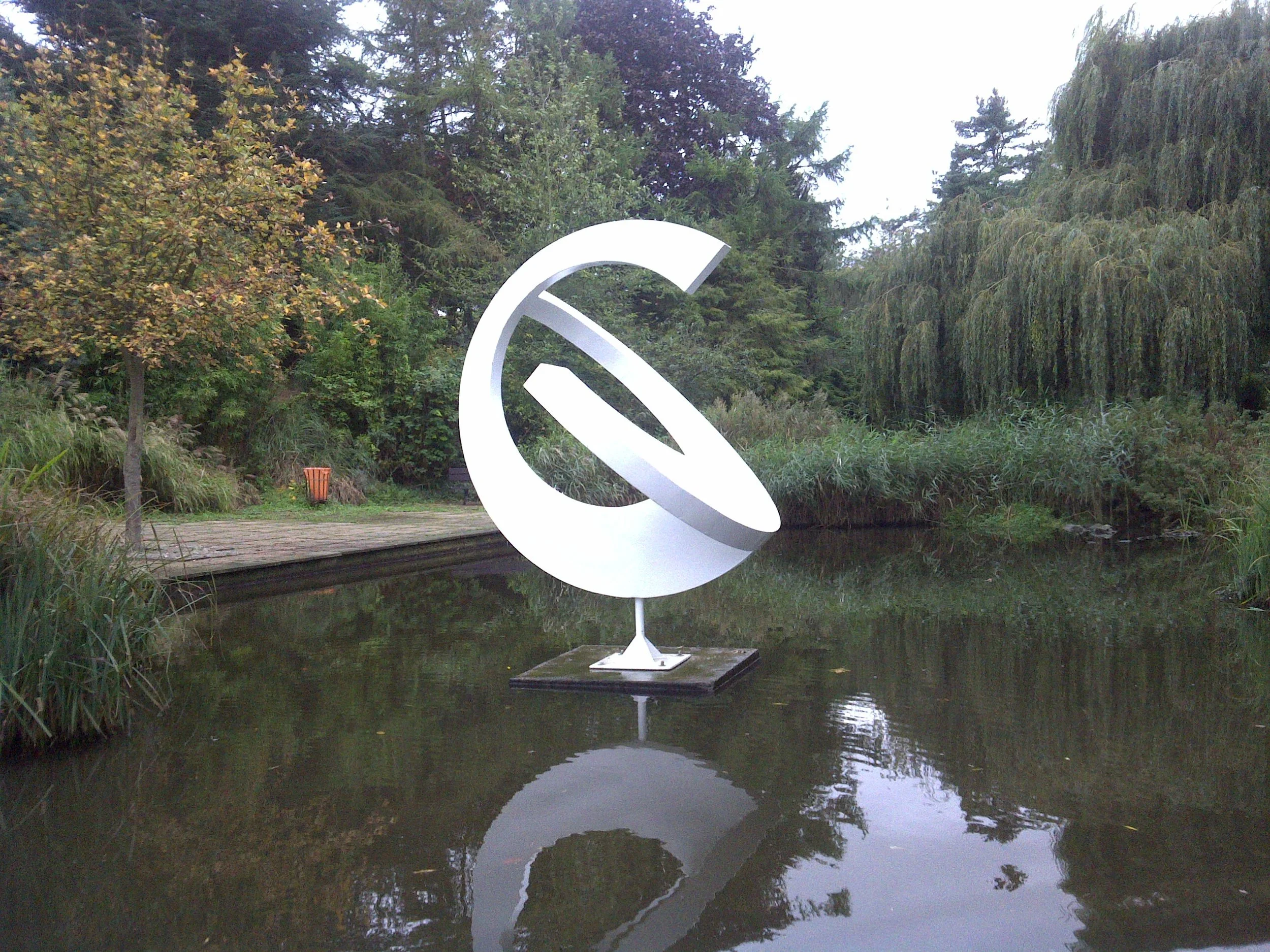 Sculpture resembling a spherical letter 'G' with an inner cutout, mounted on a platform in a pond, surrounded by trees and shrubs.