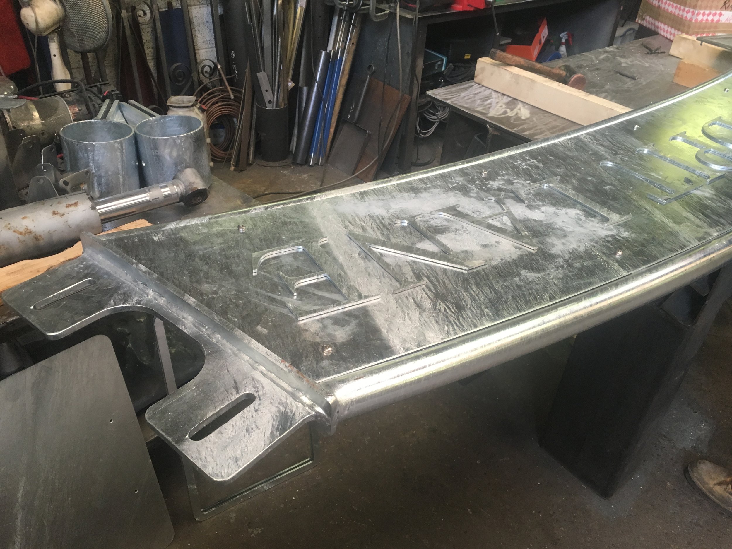 Metalwork shop with a large metallic sign reading 'RAVEN' in progress on a workbench.