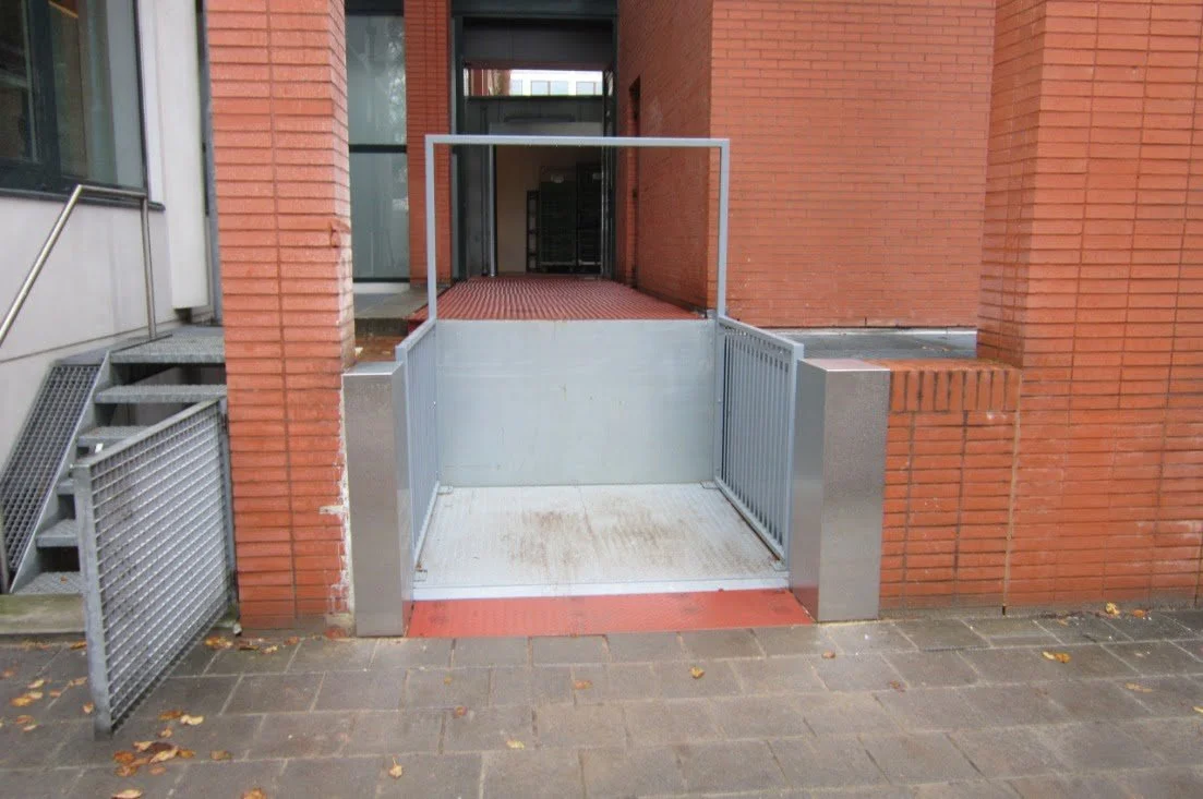 An outdoor wheelchair ramp with metal railings, leading to an entrance of a brick building, with a small set of stairs to the side.