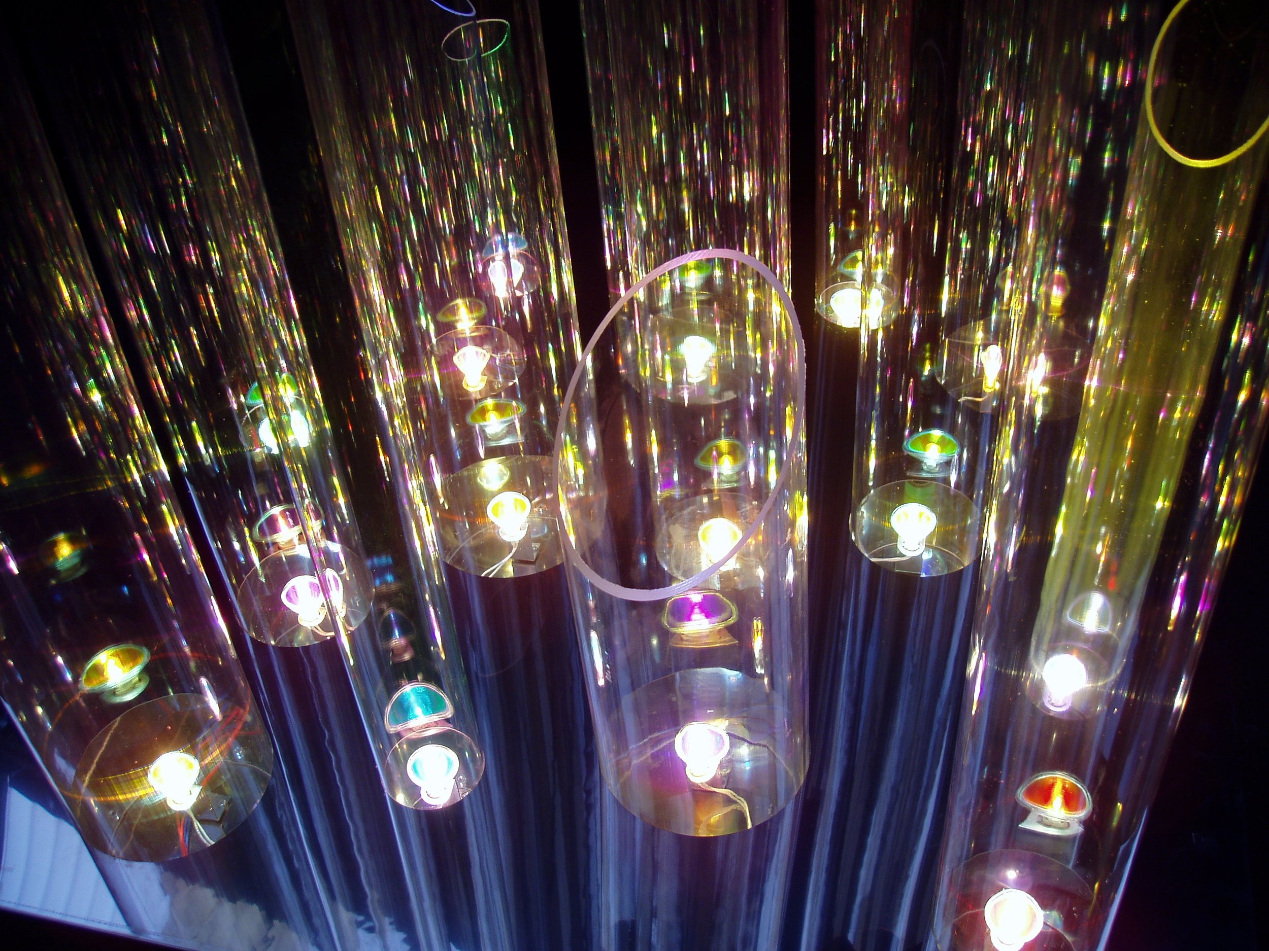 A colorful light display featuring multiple hanging light bulbs inside cylindrical transparent covers, creating a vibrant, reflective, and glowing environment.