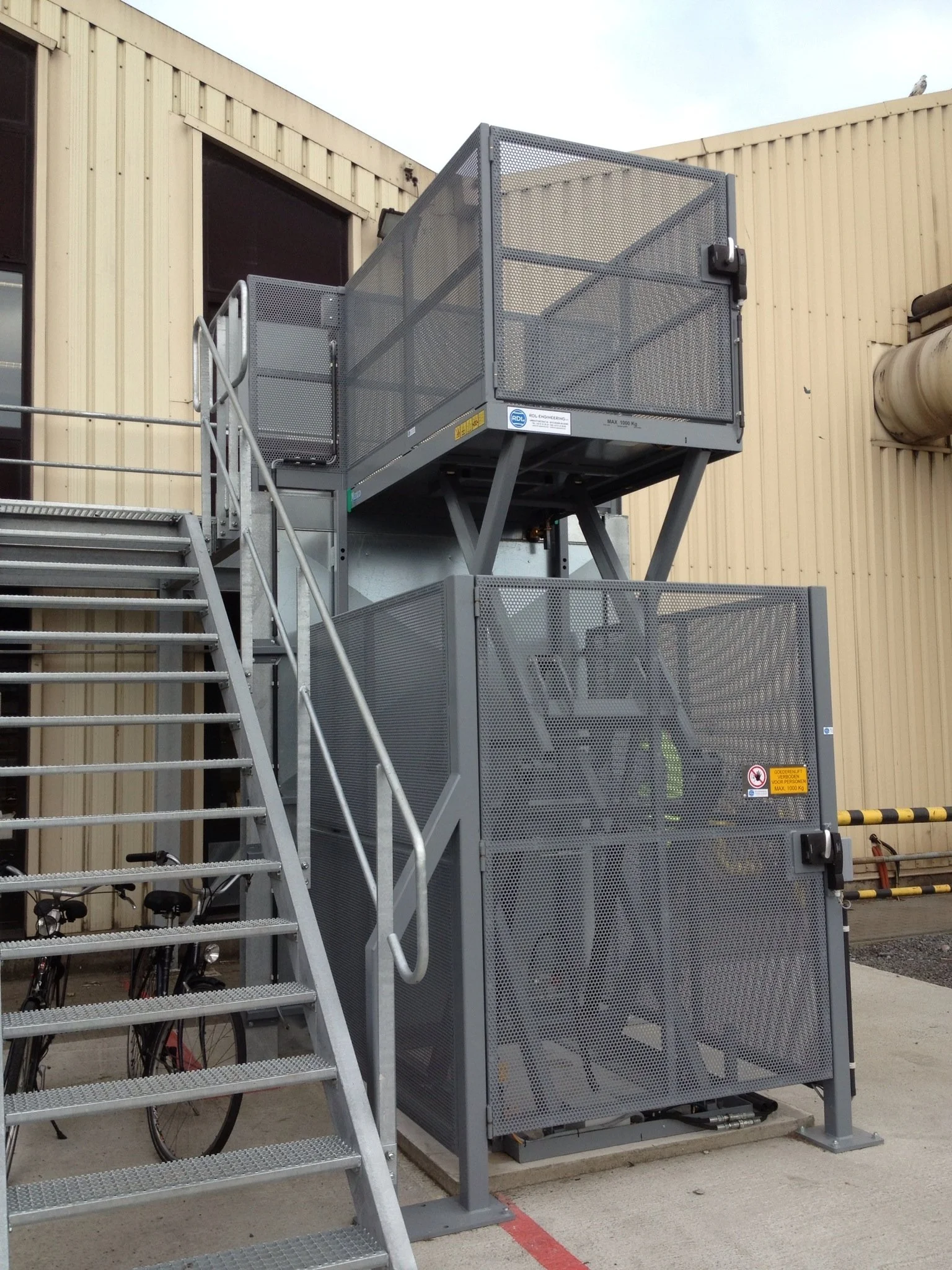Outdoor industrial lift with stairs and two enclosed compartments, adjacent to a beige metal building, with bicycles nearby.