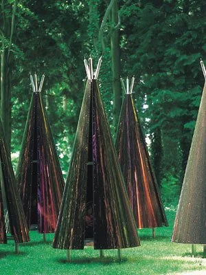 Multiple tall pumpkin-shaped sculptures with shiny dark surfaces and pointed tops outdoors on grass, surrounded by green trees.