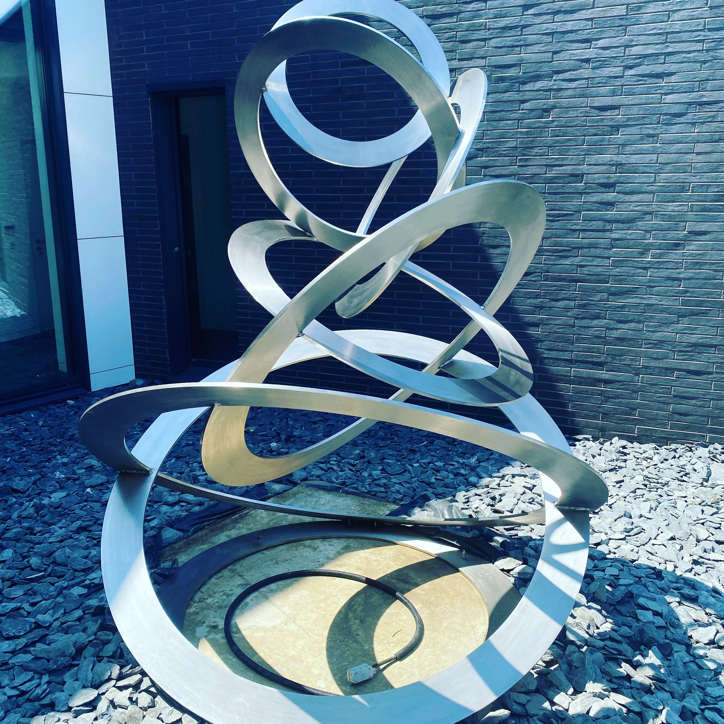 Abstract metallic sculpture with swirling loops, placed on a bed of gray rocks outside a modern building with brick and glass facade.