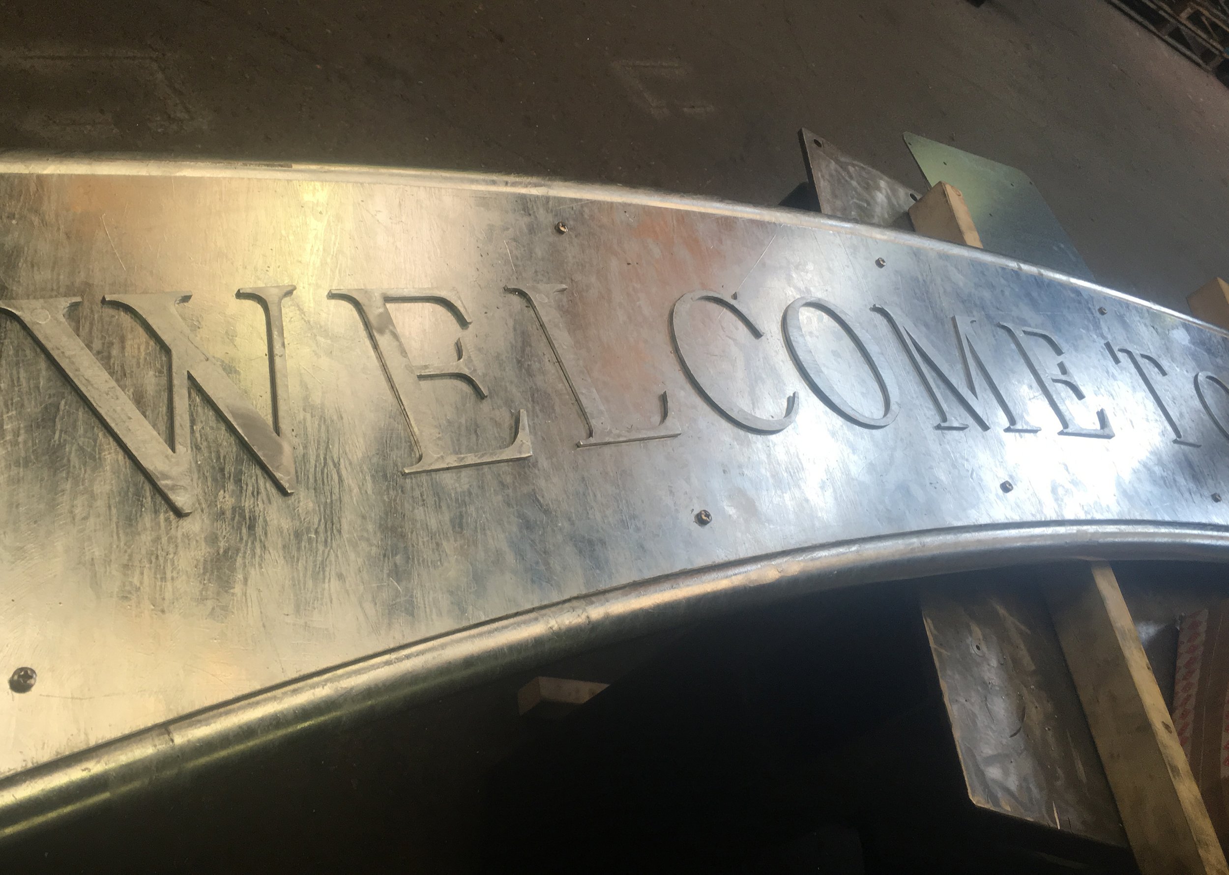 A close-up of a metal sign with the words 'WELCOMING' engraved into it, placed on a dark surface with some wooden blocks and tools around.