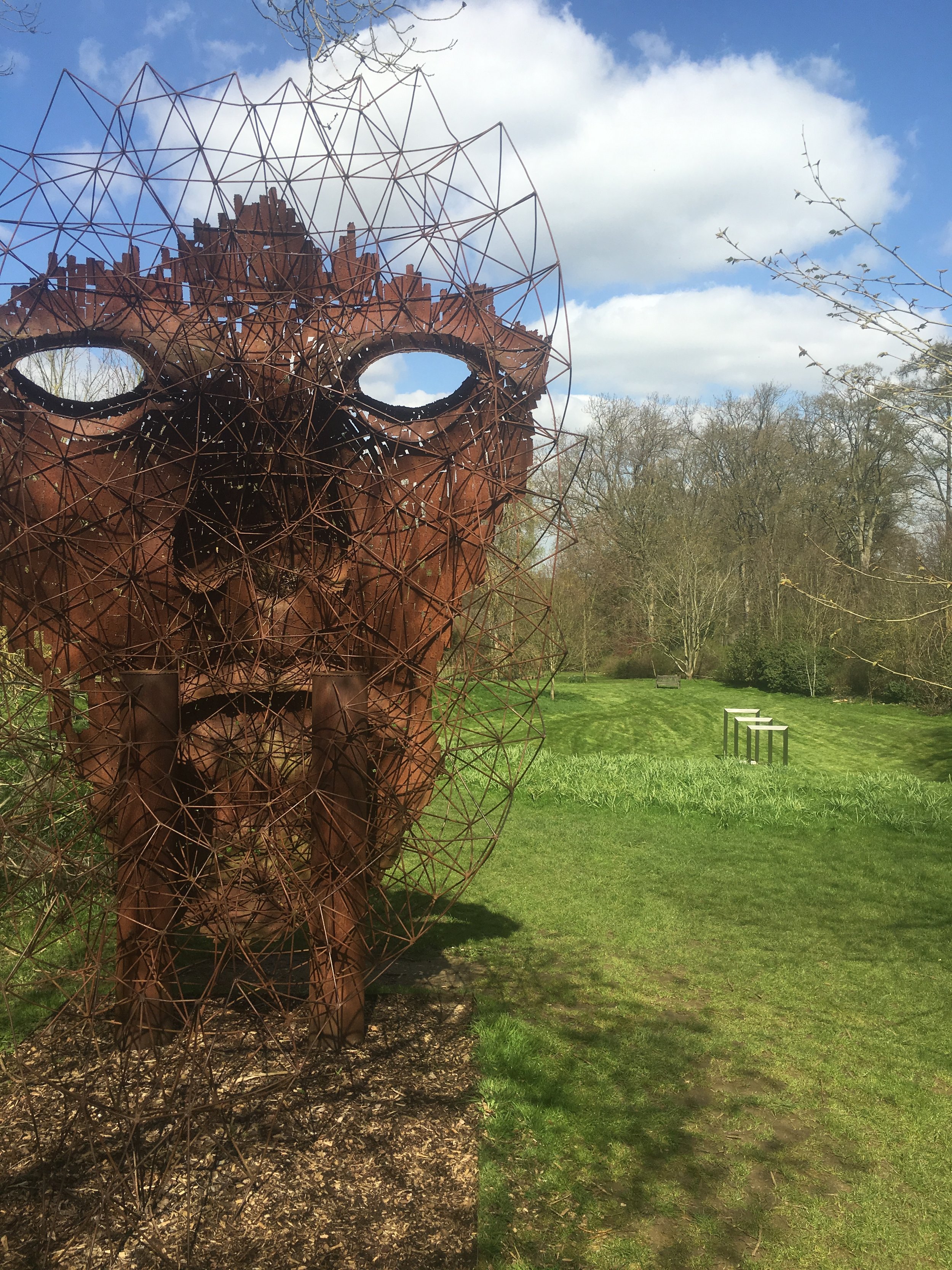 An abstract metal sculpture of a face with large eyes and a nose, made with a network of rust-colored wires, set in a park with green grass and trees in the background, under a partly cloudy sky.