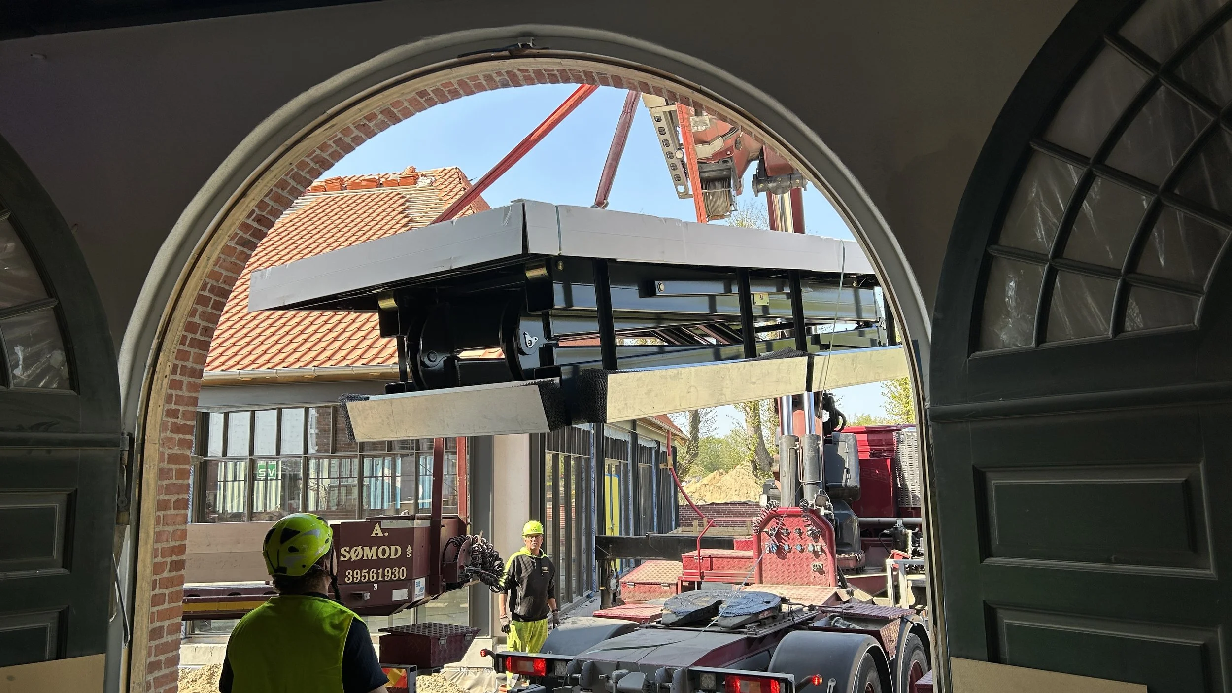Construction workers in yellow safety vests and helmets are installing a large white and black structure on a red flatbed truck outside a brick building with arched windows, under a blue sky.
