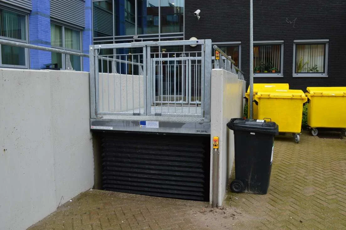 An outdoor elevator with a metal gate, built into a concrete wall, near yellow recycling bins and a black trash bin in front of a modern building.