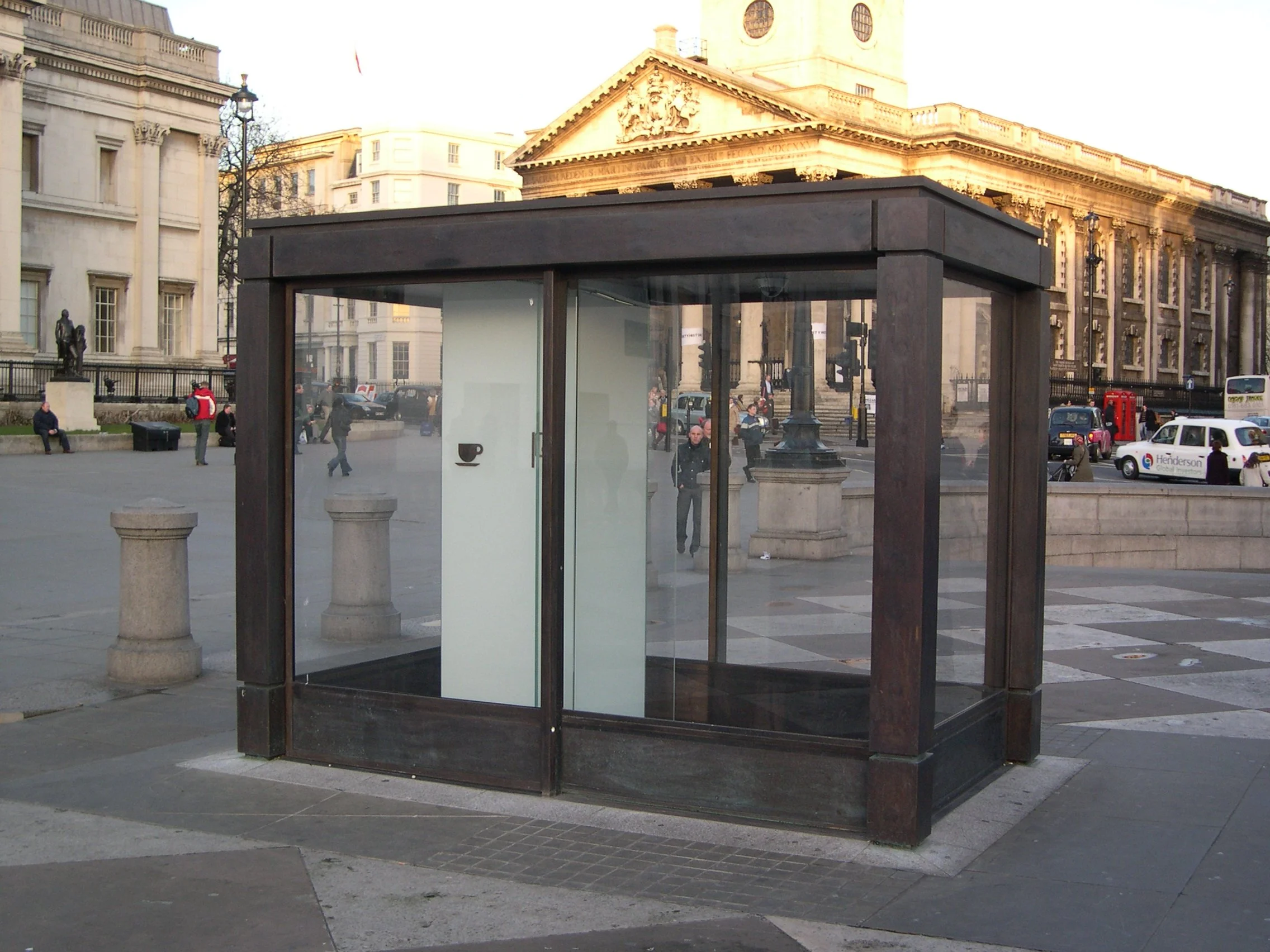 A modern glass and wood phone booth in a city square with classical architecture in the background.