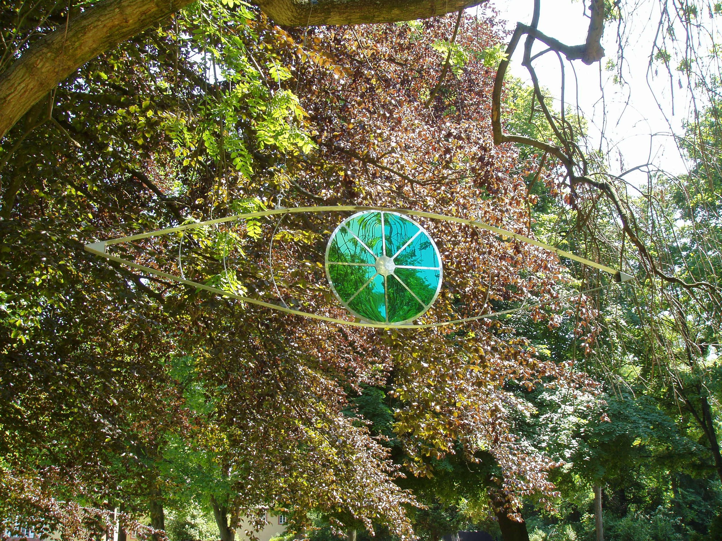 Outdoor garden scene with a large, decorative hanging piece resembling an eye with green and blue stained glass, surrounded by trees with green and reddish-brown leaves.