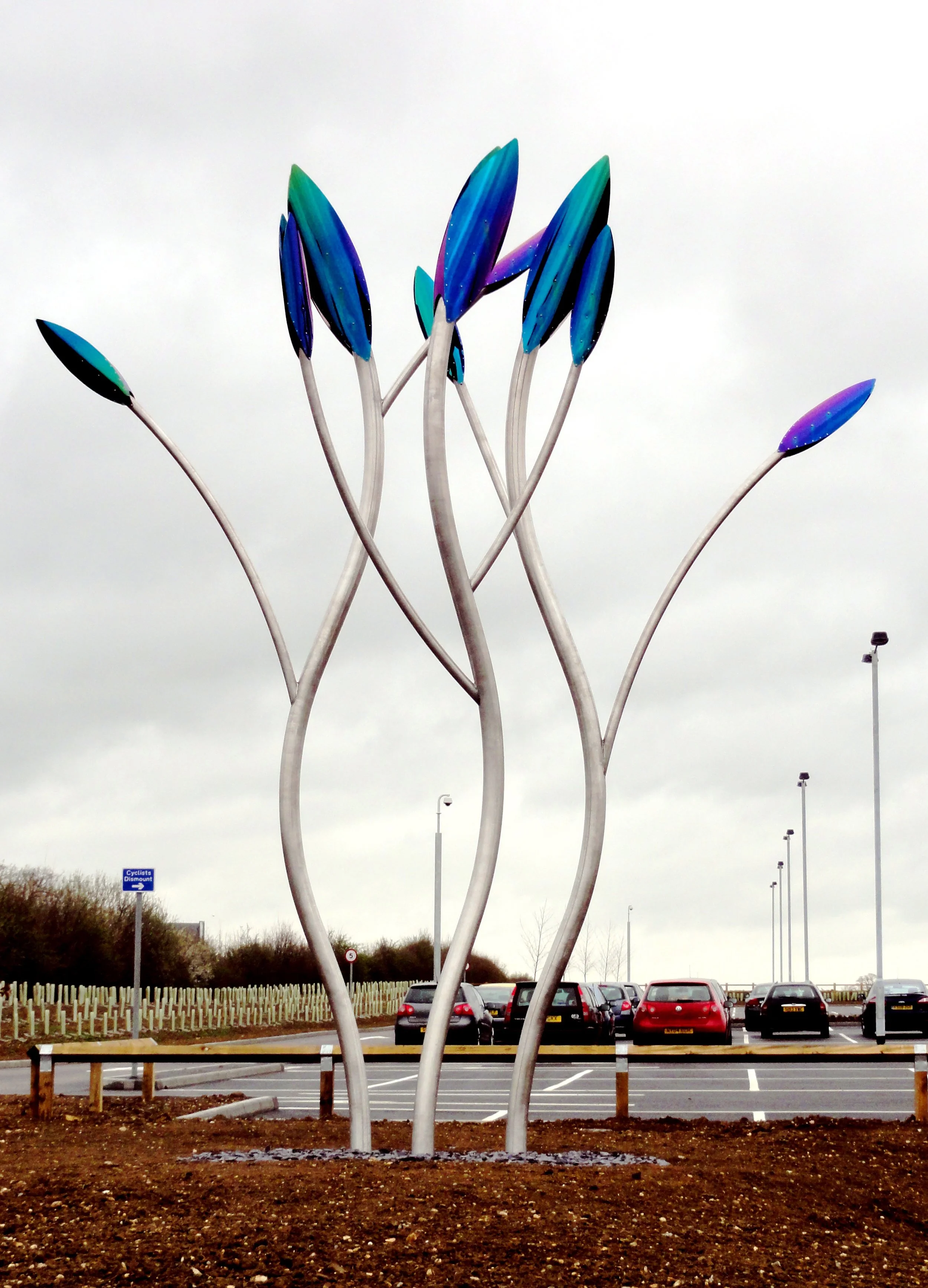 Colorful metal sculpture resembling a bouquet of tulips in a parking lot with cars and lamp posts in the background.