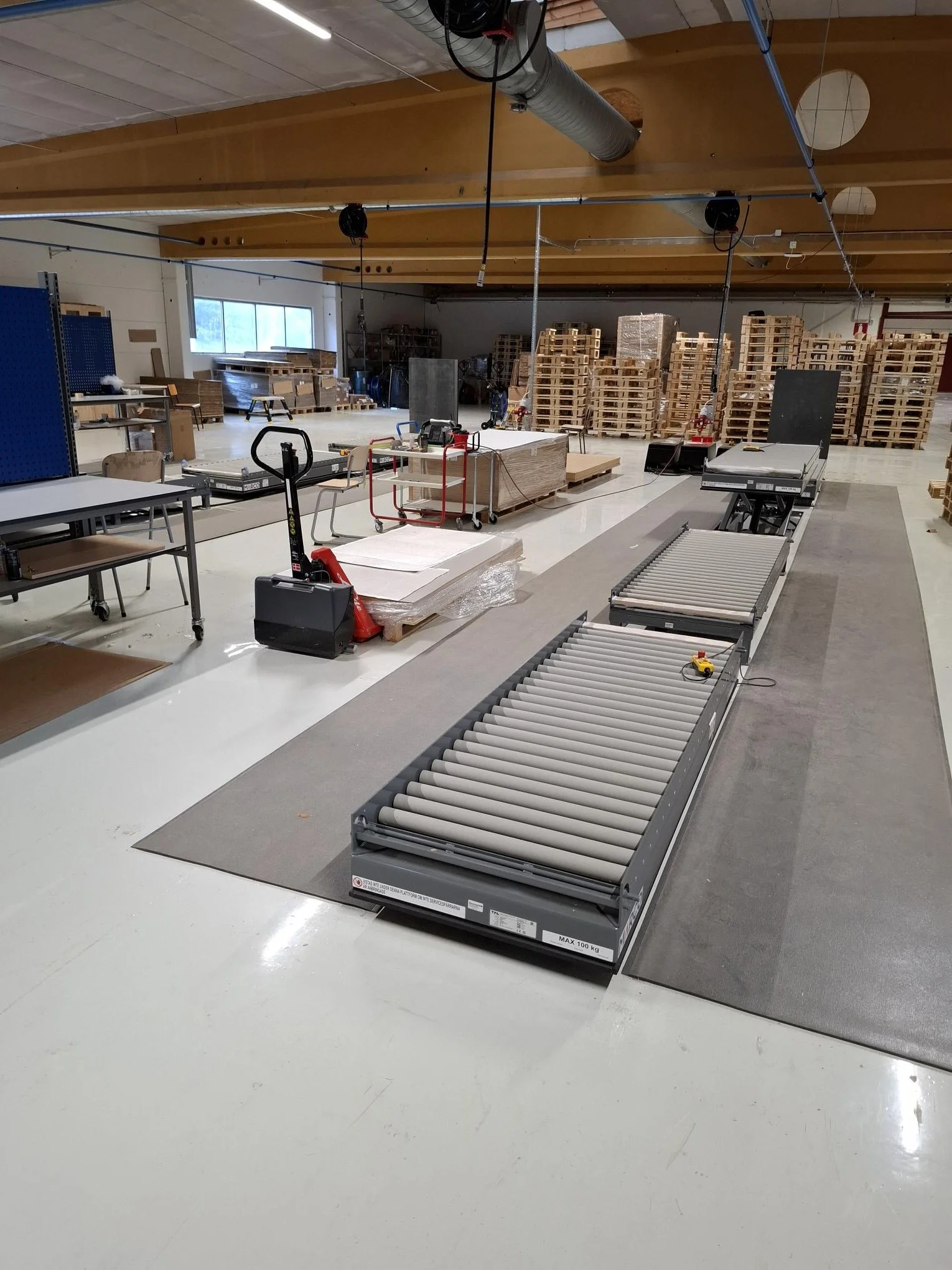 Warehouse or factory space with conveyor rollers, tools, pallets, and stacked wooden pallets in the background.