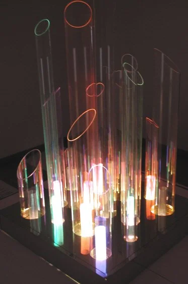 Colorful, illuminated glass tubes of various heights and shapes arranged on a dark surface.