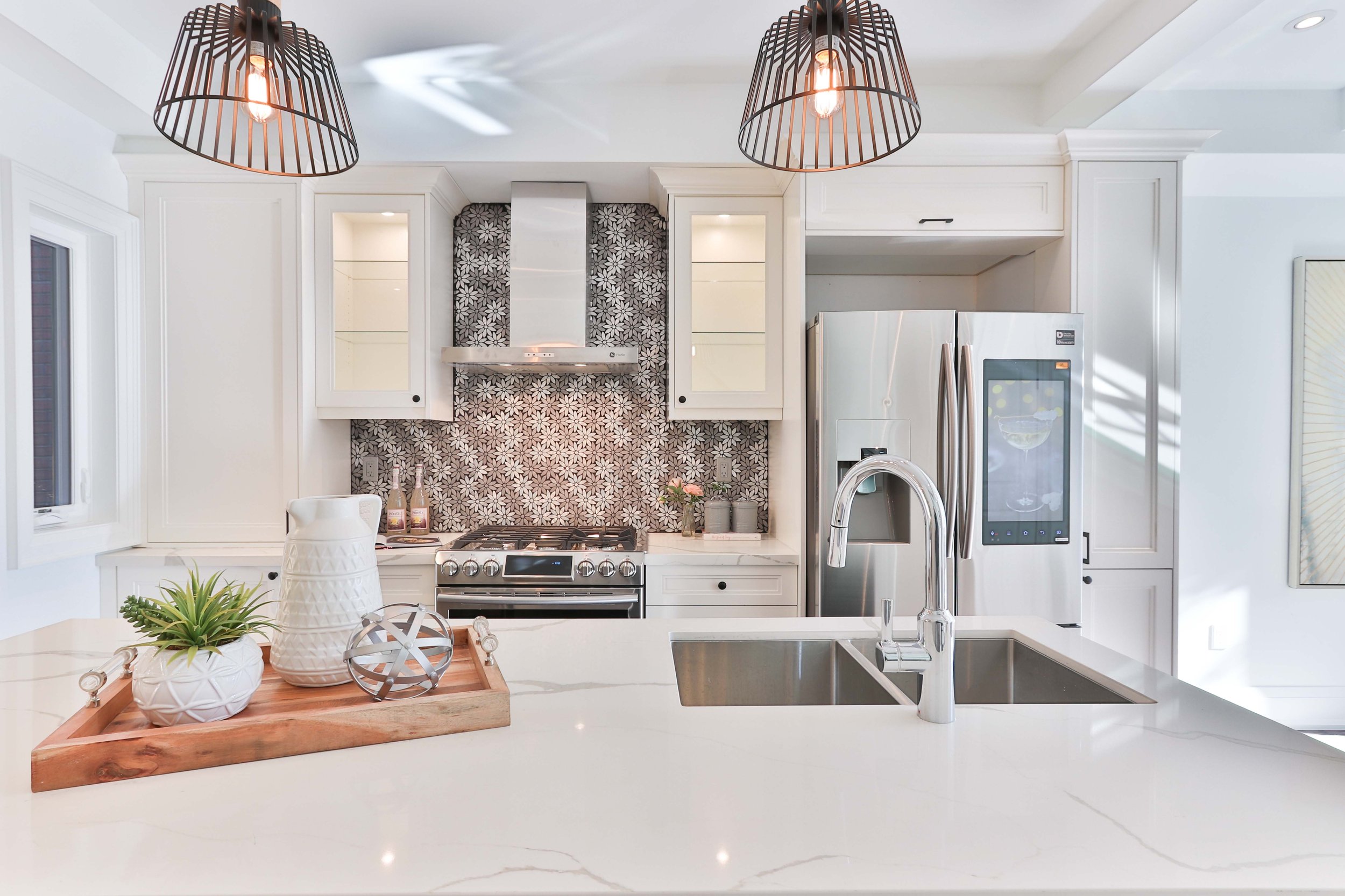 Modern kitchen with white cabinets, stainless steel appliances, patterned backsplash, pendant lights, plants and decorative items on the counter