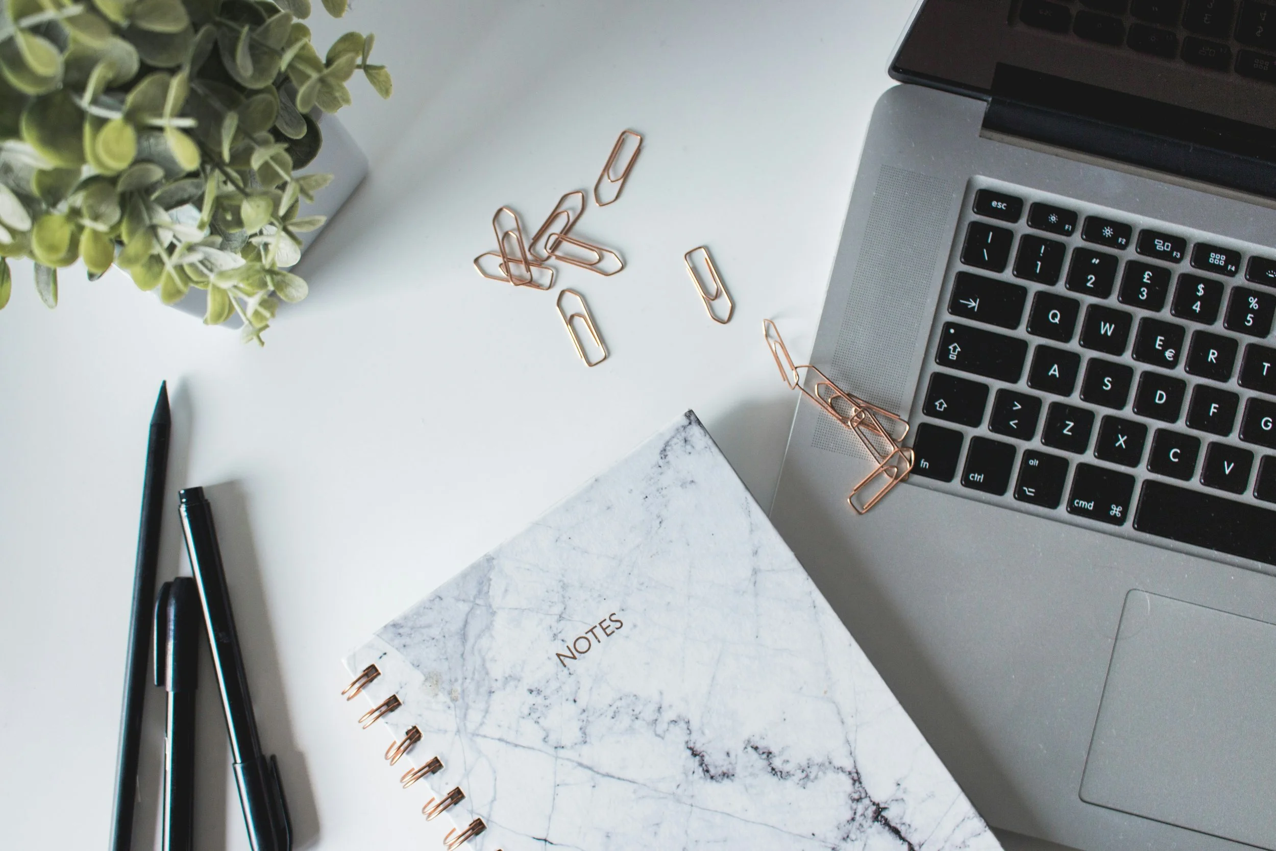 A workspace with a laptop, a notebook labeled 'Notes,' pens, paper clips, a plant, and a notebook on a white desk.