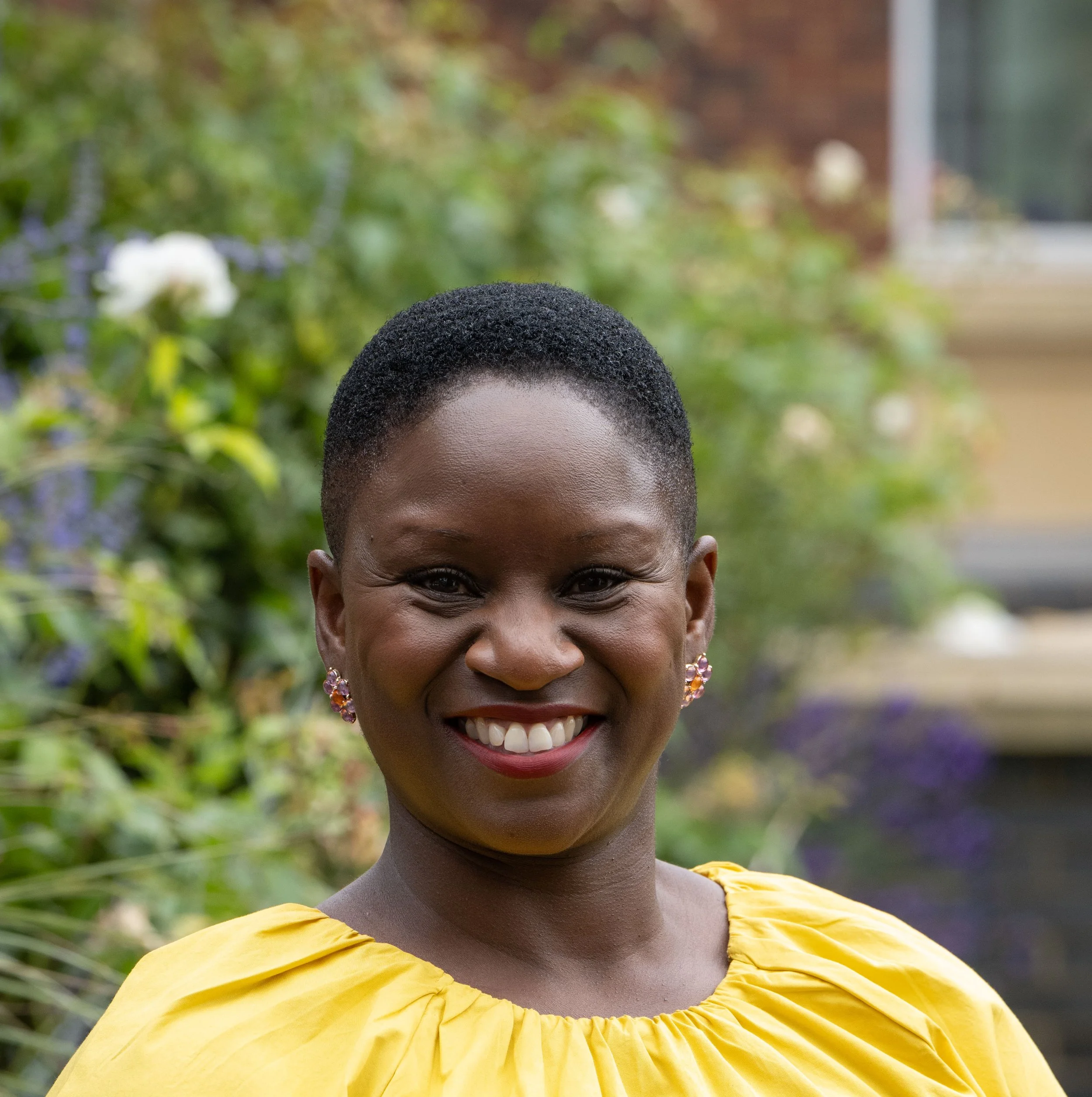 A woman with very short, textured hair smiling outdoors, wearing a yellow top and colorful earrings, with green plants and flowers in the background.
