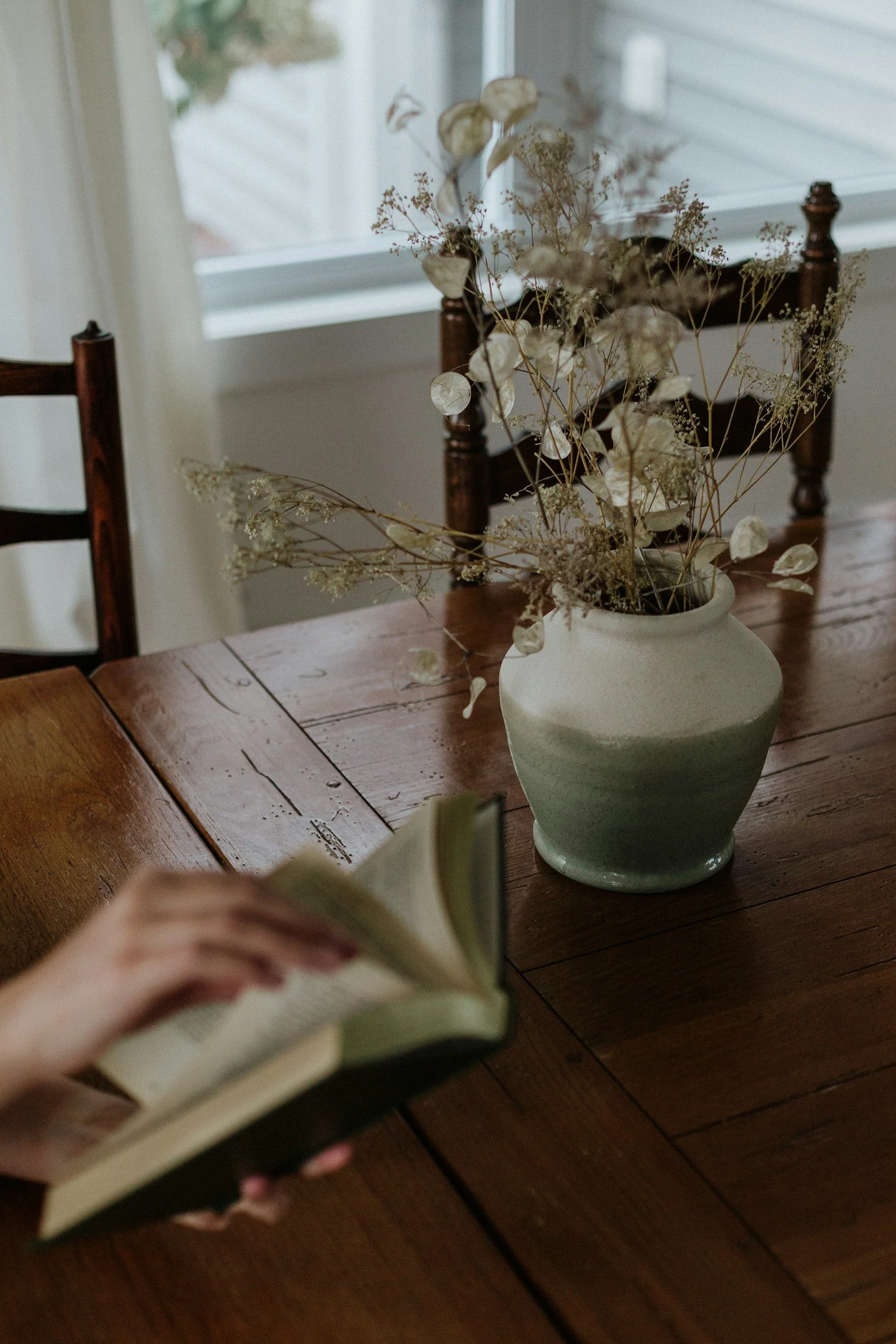 In the foreground a person is holding a book reading while sitting at a writers desk with a vase of flowers on it