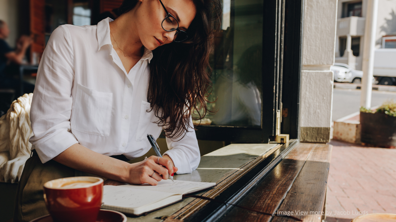 A woman with long dark hair and glasses wearing a white shirt is sitting at a wooden table, writing in a notebook with a pen, next to a cup of coffee in a cozy cafe.