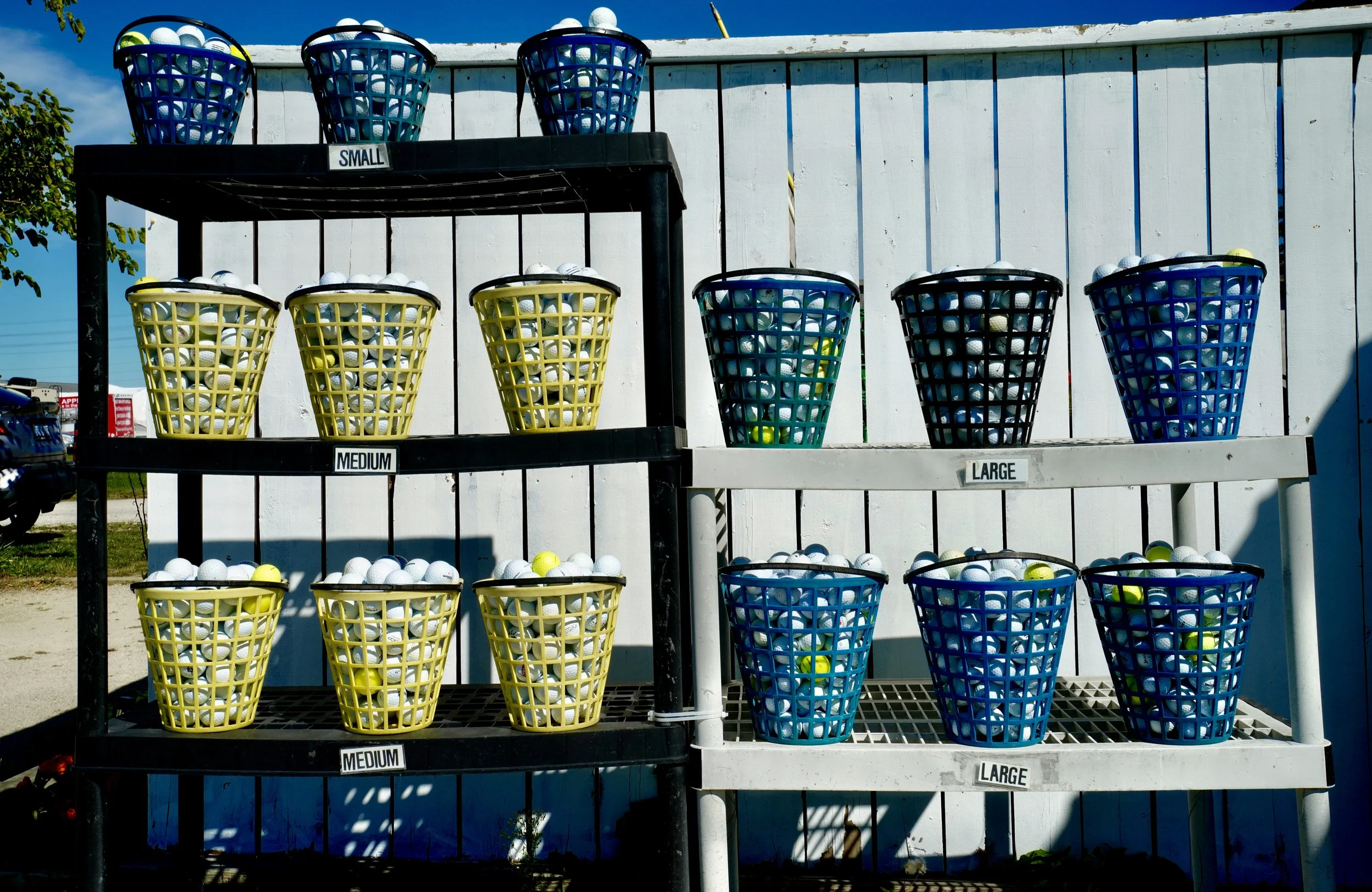 Buckets of golf balls arranged on outdoor shelves, labeled by size: small, medium, and large, with a white wooden fence in the background.