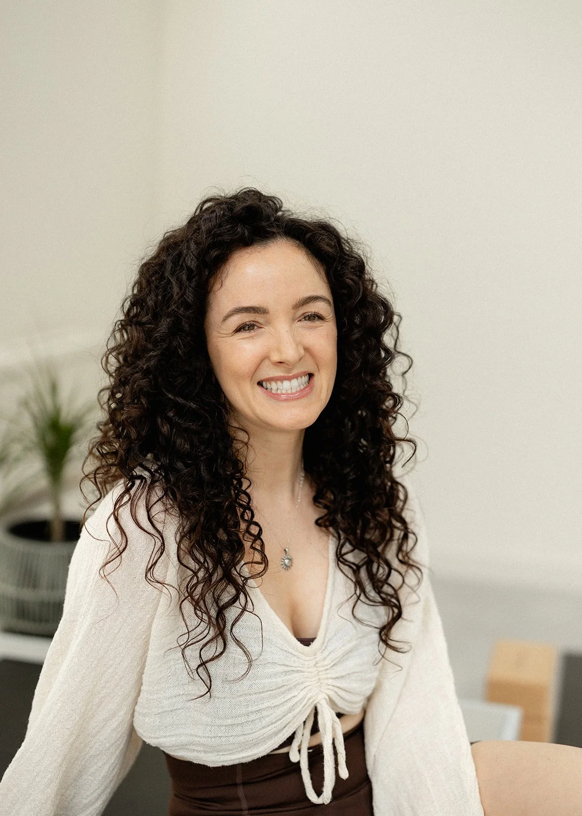 A woman with curly hair sitting cross-legged on a bed by a window with a scenic view of a sunset over a rural landscape.