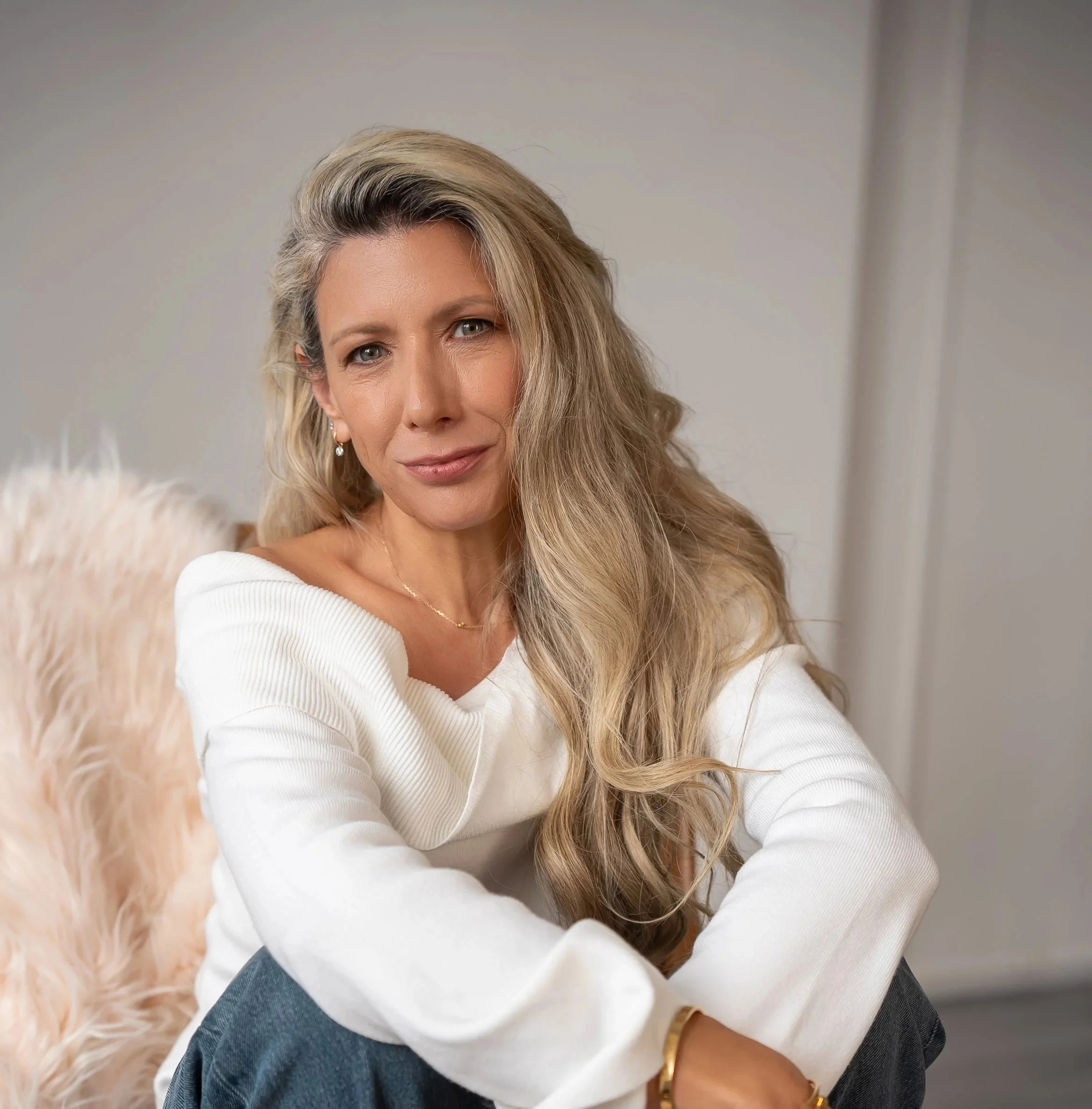 A woman with long blonde hair, wearing a white off-shoulder top and sitting on a chair with a furry pillow behind her.