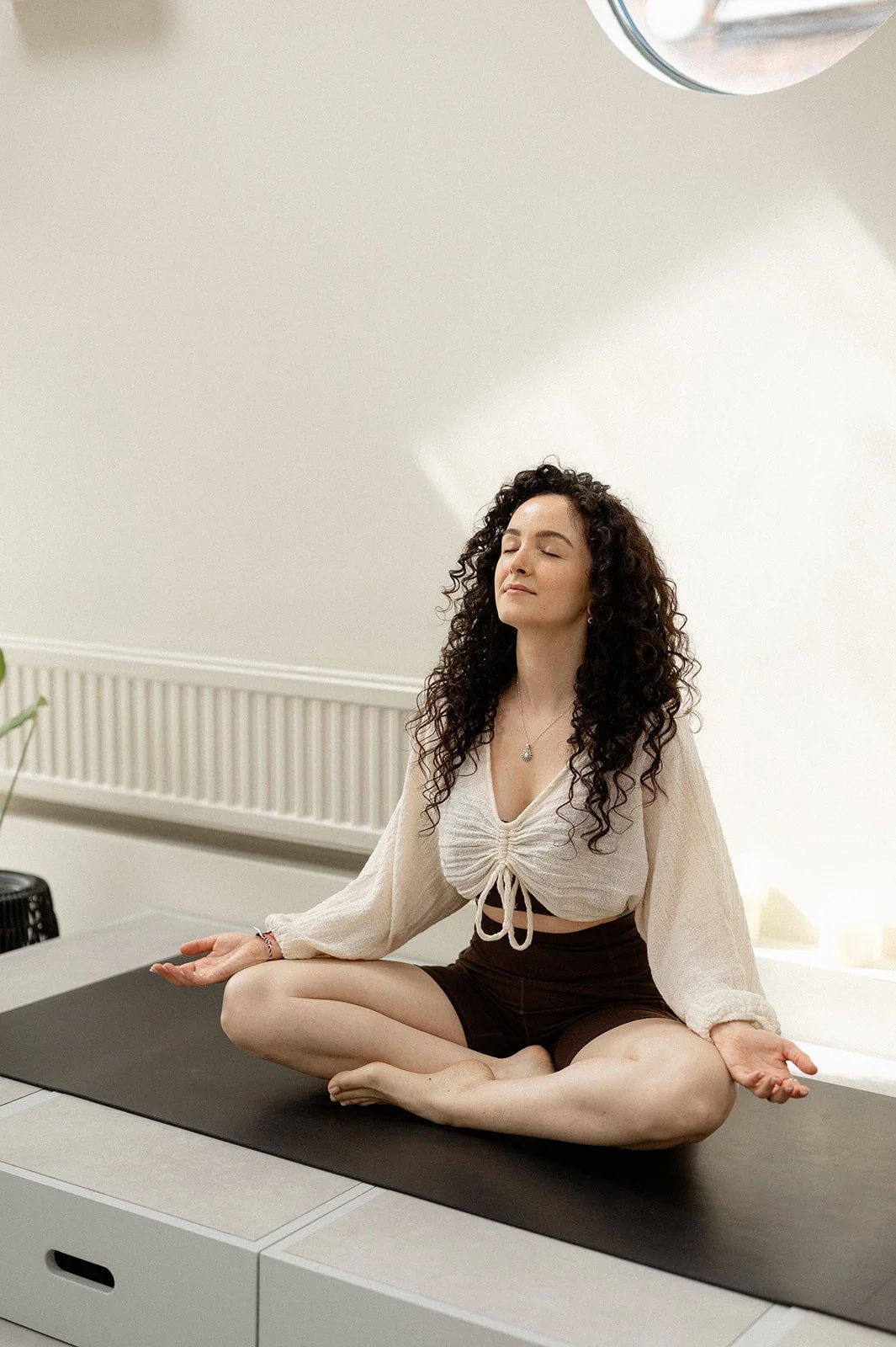 A woman with curly hair sitting cross-legged on a bed by a window with a scenic view of a sunset over a rural landscape.