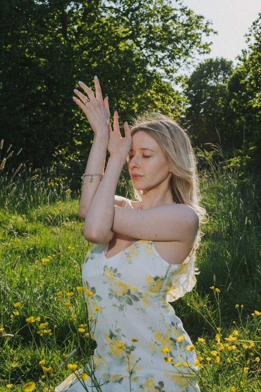 A young woman with blonde hair in a floral dress practicing yoga outdoors in a green field surrounded by trees, with sunlight filtering through.