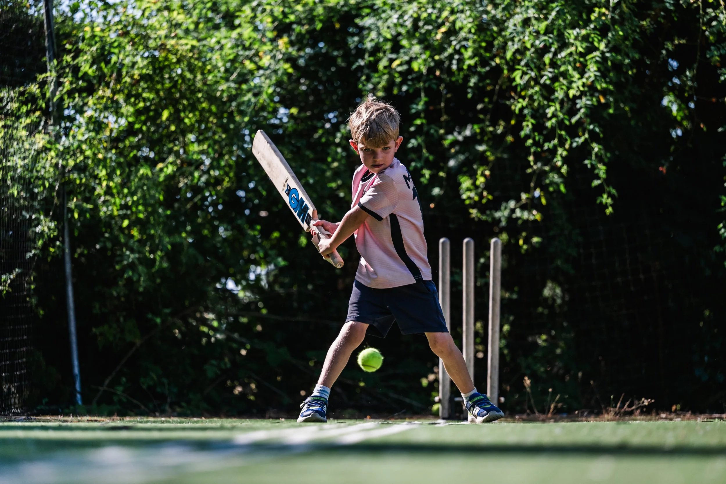 A young boy playing cricket outdoors, swinging a cricket bat, with a cricket ball and wickets in the background.