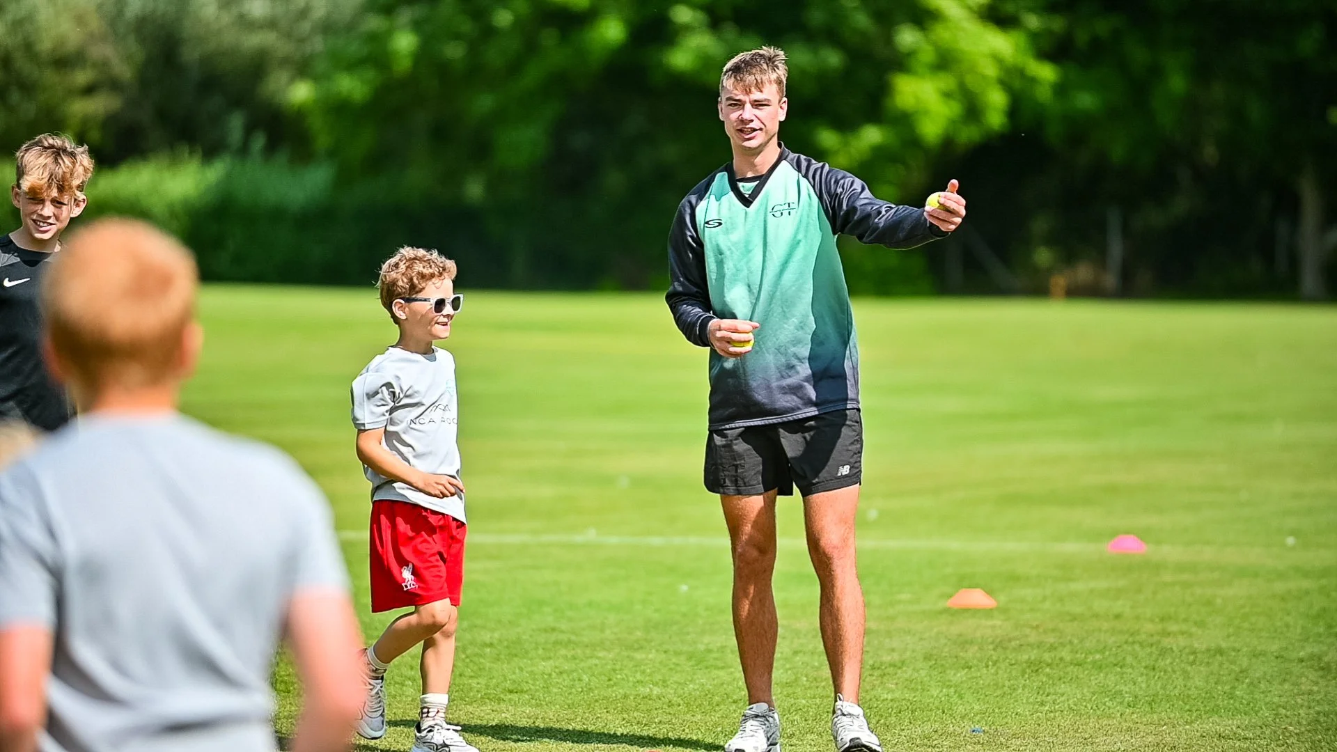 A group of children and an adult coach on a grassy field, engaging in a sports activity on a sunny day with trees in the background.