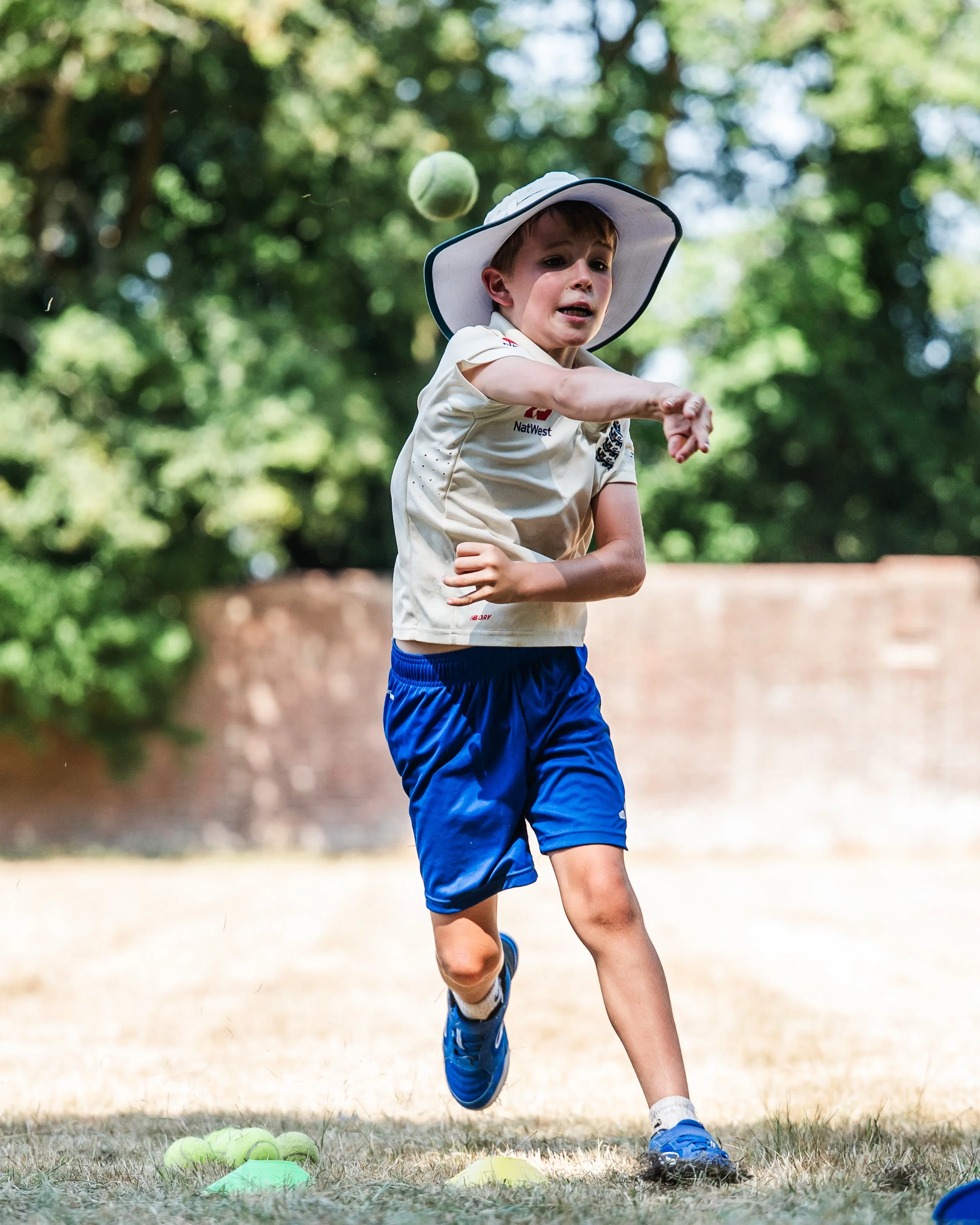 Young boy in sports uniform running in a park, with tennis balls and cones on the ground, during daytime.