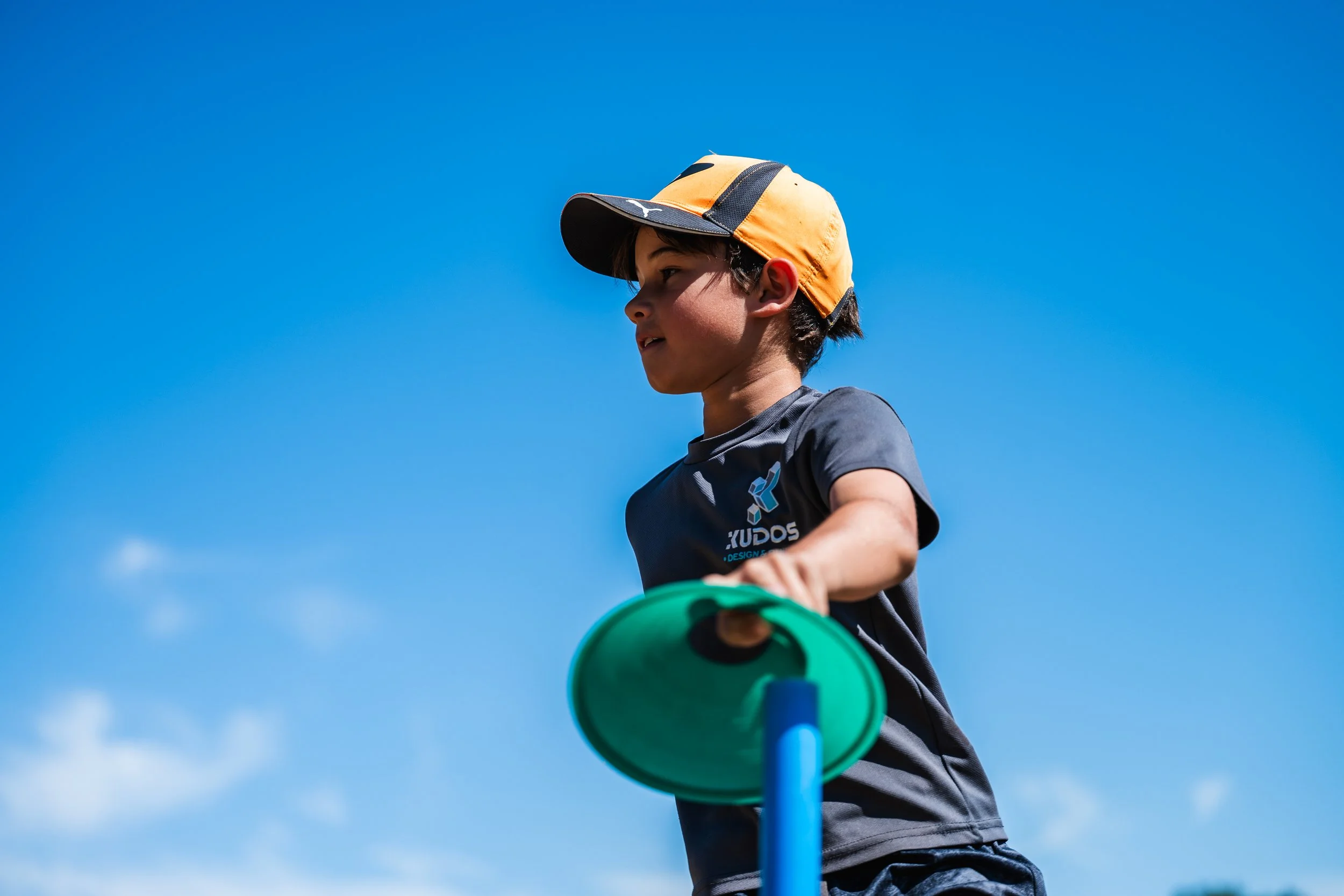 A boy wearing a cap and athletic clothing, reaching out towards a green disc while playing outside under a clear blue sky.