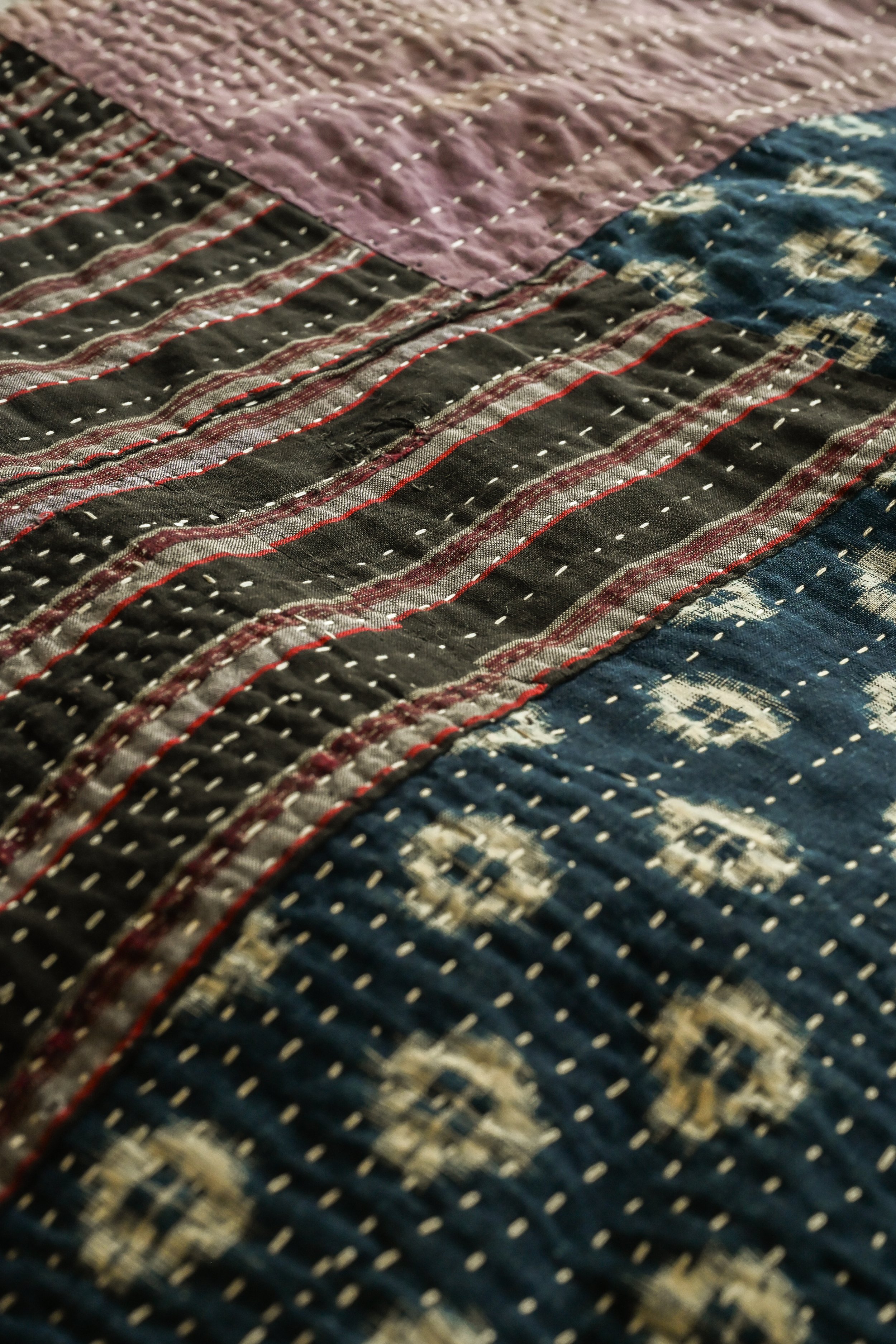 Close-up of a multicolored quilt with detailed stitching, featuring sections of purple, black with red and white lines, and blue with white patterns.