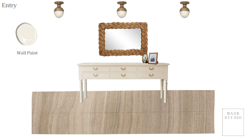 A white hallway console table with drawers, on a light wood floor, centered below a large woven frame mirror, with a framed photo and small decorative items on it. Three ceiling lights are above. To the left, there is an entry sign and a wall paint sample.