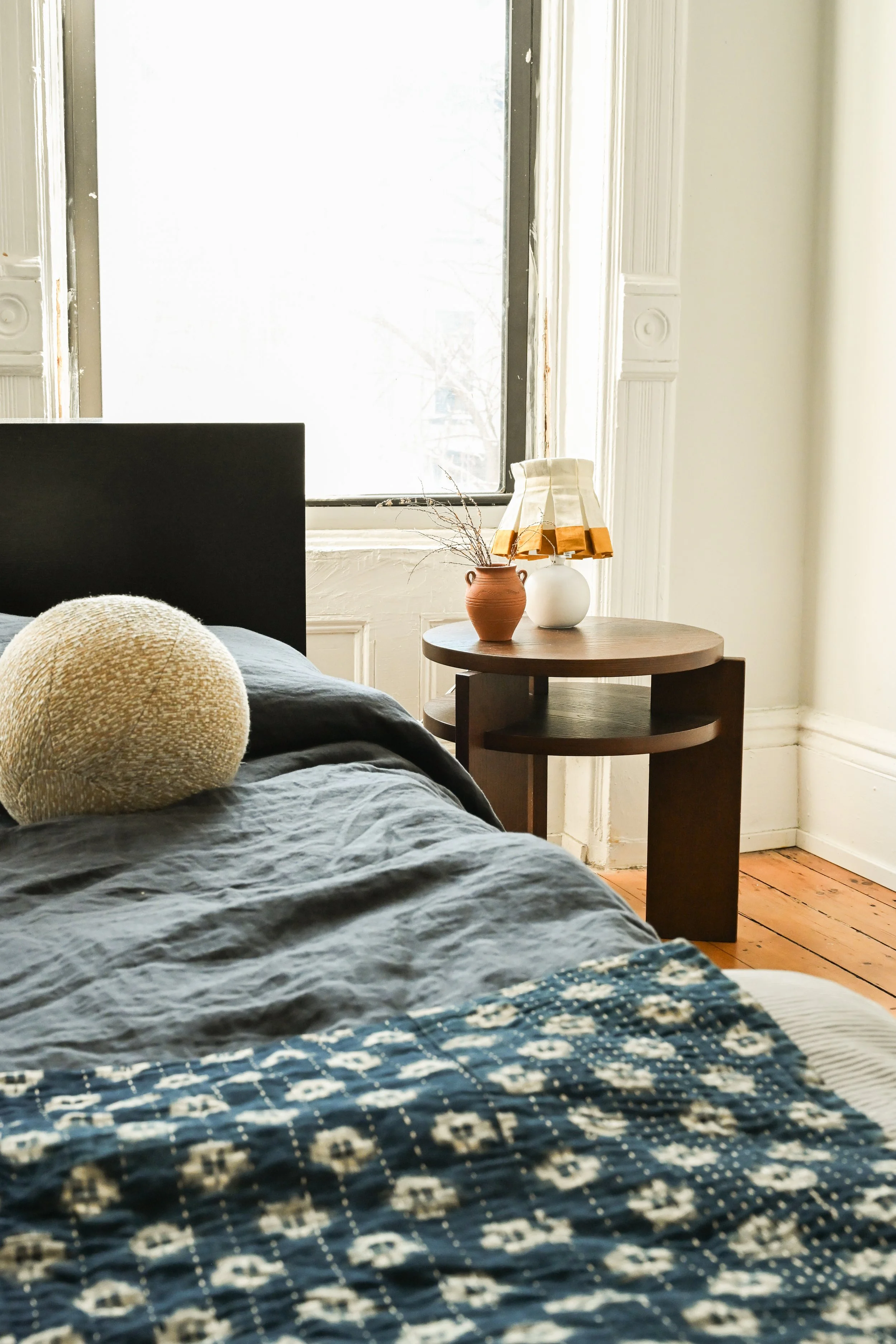 A cozy bedroom scene with a bed covered in a dark blue quilt with white patterns, a rounded textured beige pillow, a small wooden side table with a pair of vases (one terracotta and one white), and a window with white framing and a view of a tree outside.