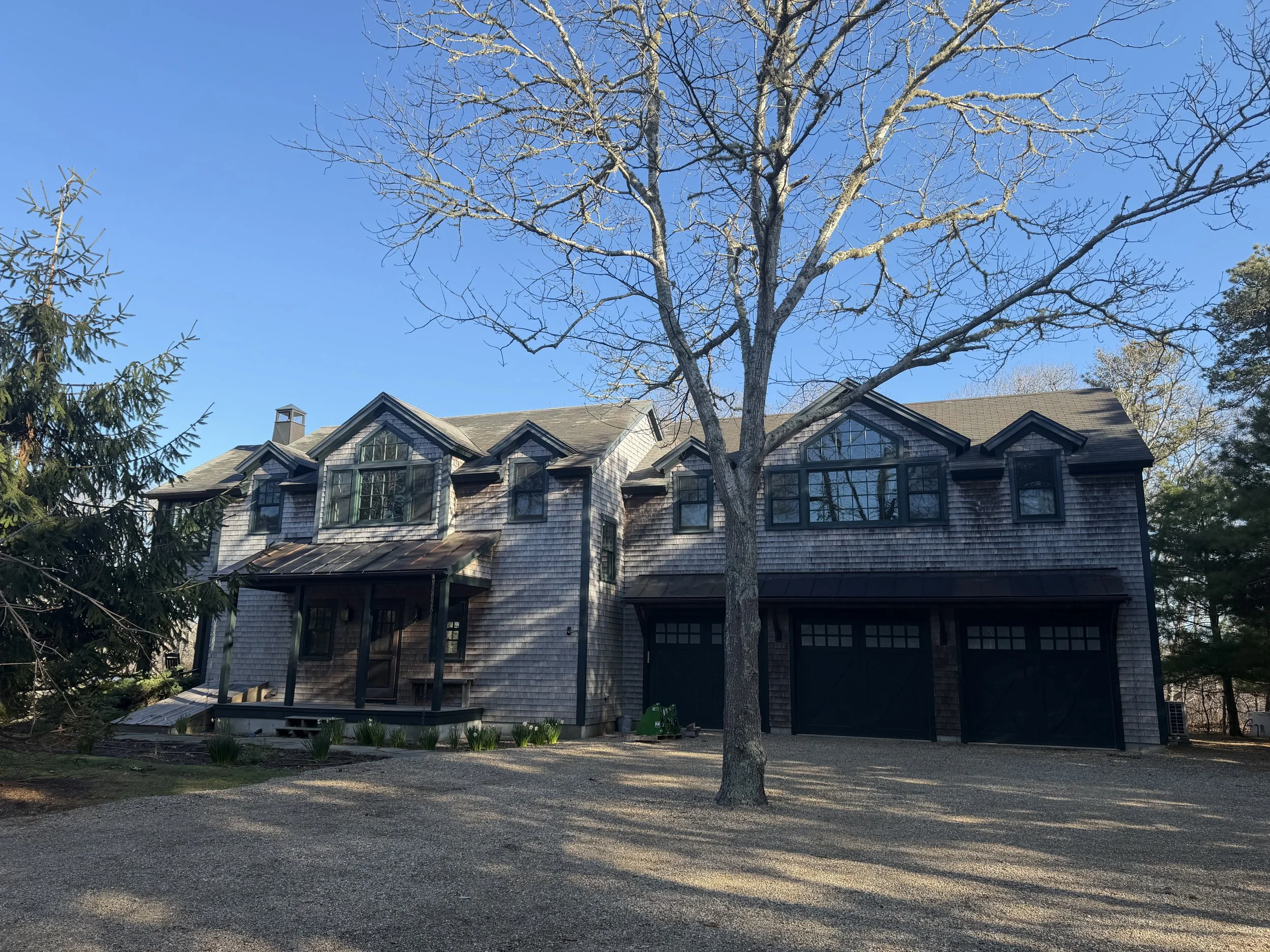 A large house with gray shingles, multiple pointed roofs, and numerous windows, surrounded by trees and a gravel driveway, under a clear blue sky.