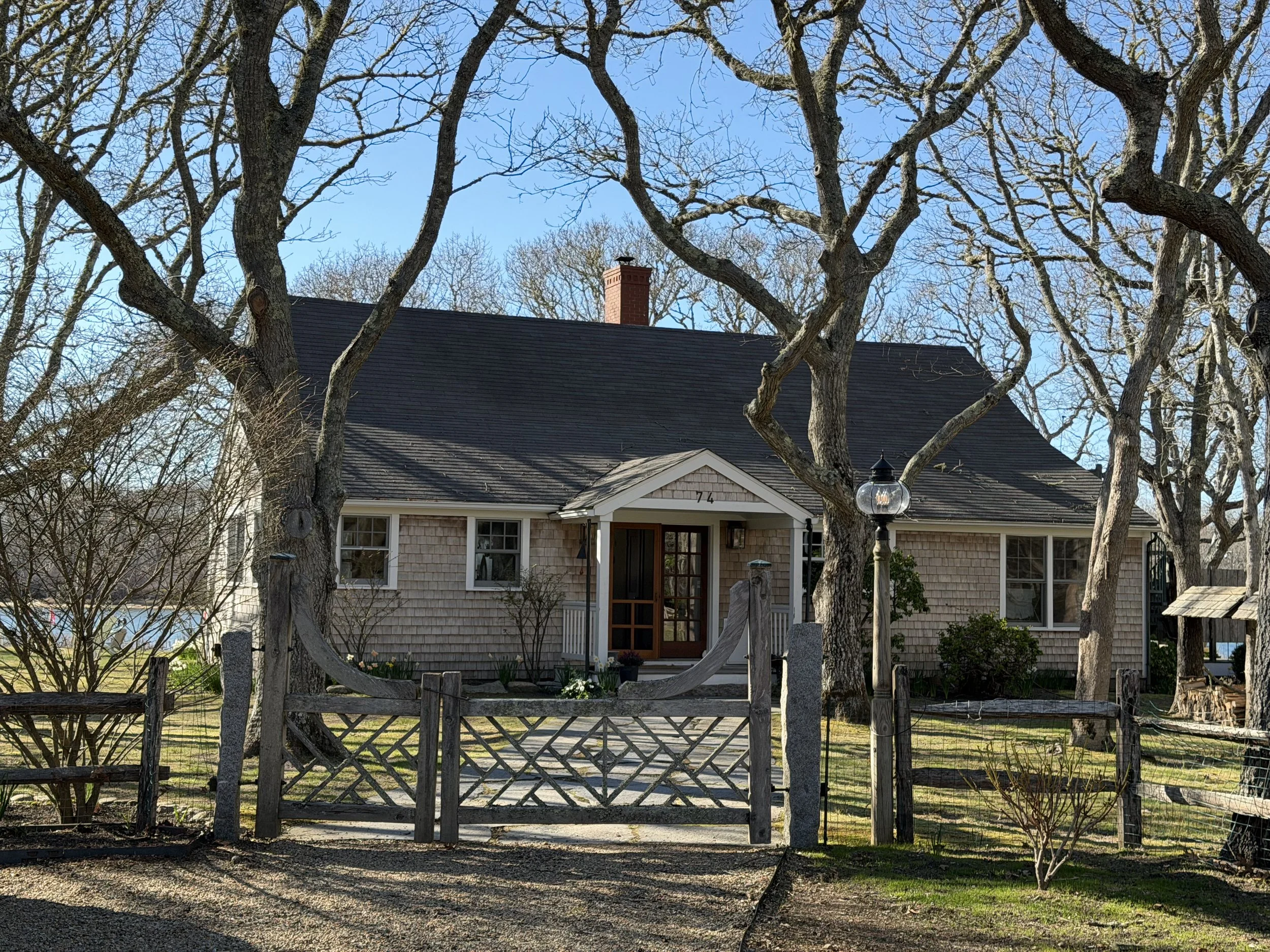 Front yard view of a house with a black roof, chimney, and beige shingle siding. There are large trees with no leaves partially obscuring the house, a wooden gate in the fence, a mailbox, and a lantern-style lamp post near the walkway.