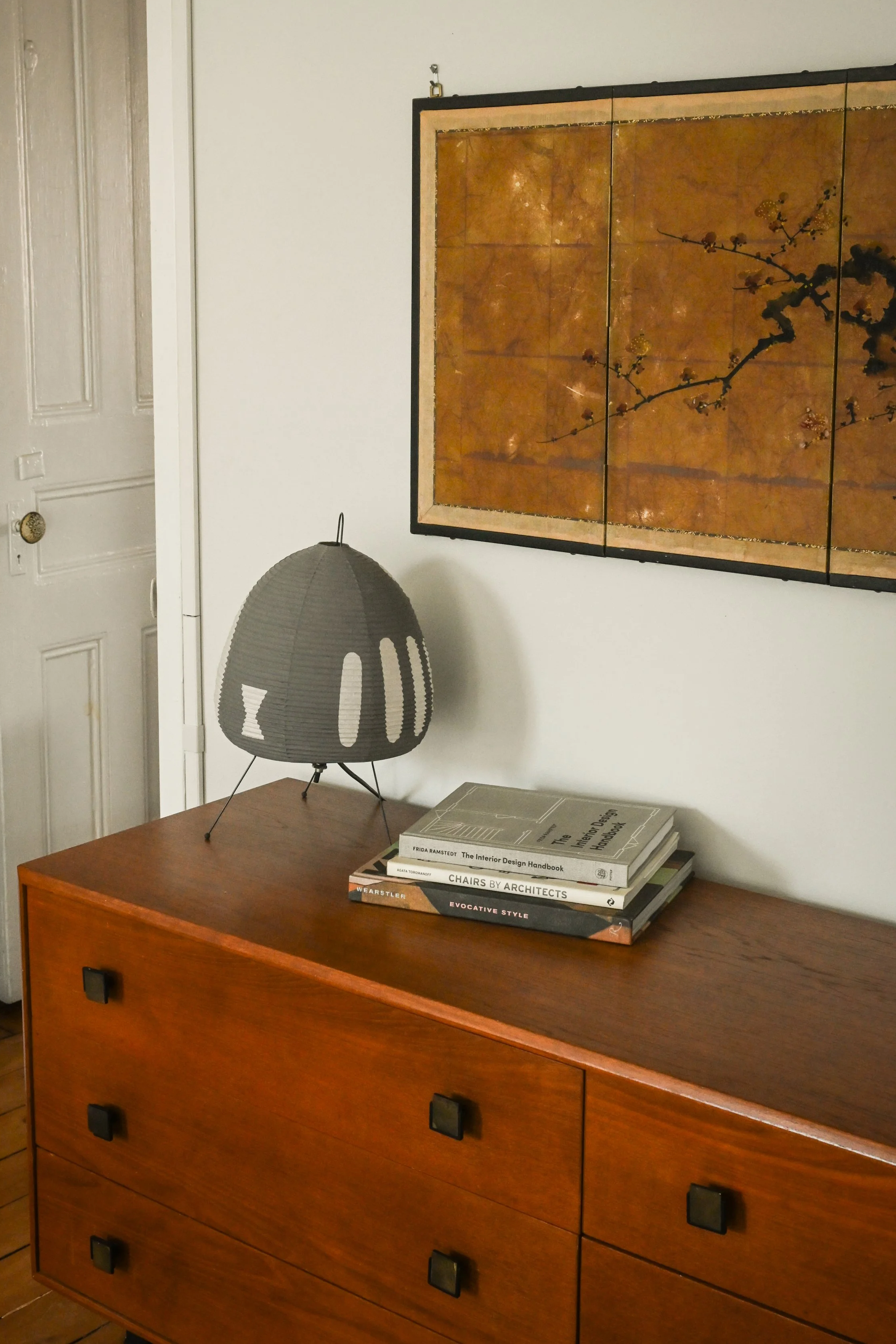 A wooden mid-century modern dresser with six drawers, black square knobs, on a wooden floor in a room with white walls. A small stack of books is on top of the dresser, including titles about interior design and architecture. A gray lamp with a conical shade and tripod base sits on the left side of the dresser, and a framed abstract artwork with warm tones and branches with blossoms hangs above the dresser.