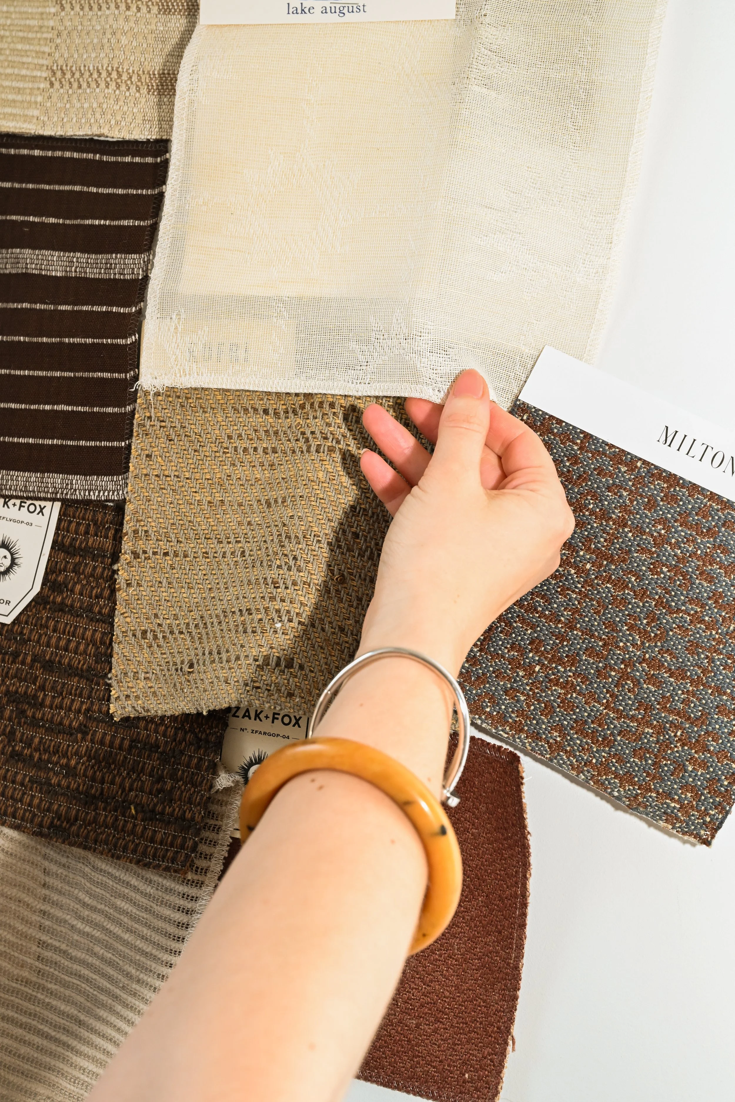 A person's hand holding a piece of fabric sample swatch. The fabric is light-colored with a mesh-like texture. Multiple fabric swatches with different patterns and textures are laid out on a surface. The person's wrist has a wooden bangle, and the background appears to be a neutral wall.