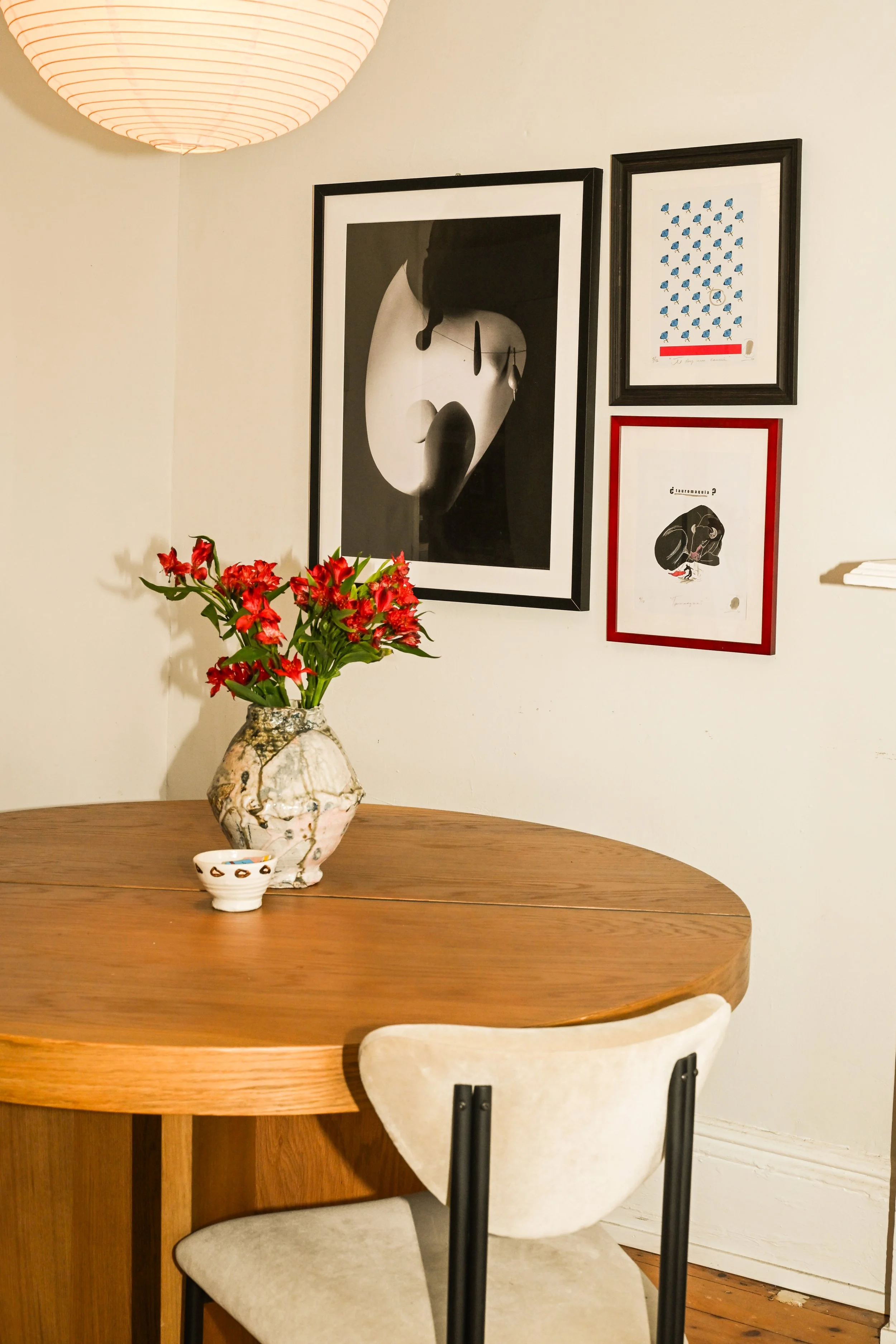A wooden dining table with a floral vase holding red flowers and a small decorative bowl on top. A white cushioned chair with black metal legs is positioned at the table. On the wall behind, there are four framed art pieces, including abstract black and white images, a blue pattern, and a black and white illustration. A paper lantern light fixture hangs above.
