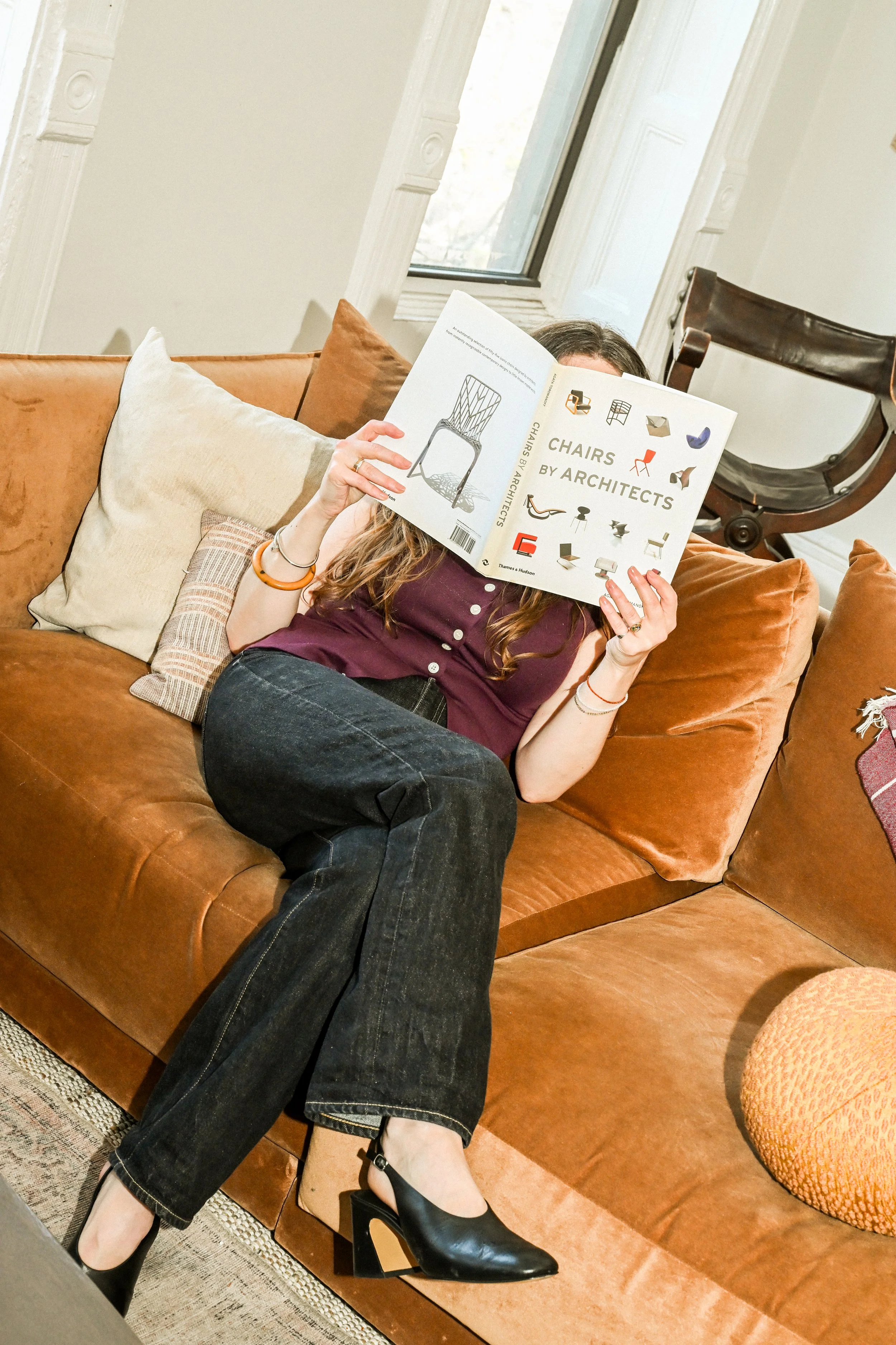 A woman sitting on a brown leather sofa, reading a book titled 'Chairs by Architects' in a living room with a large window and various pillows, wearing black high heel shoes and black jeans.