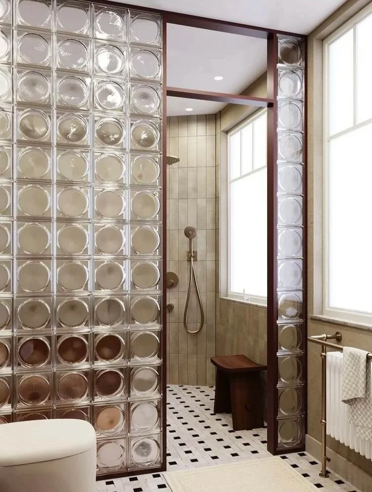 Bathroom with a curved shower area, glass block wall partitions, and a bench in the shower. Large windows letting in natural light, with beige tiles and black and white checkered floor.